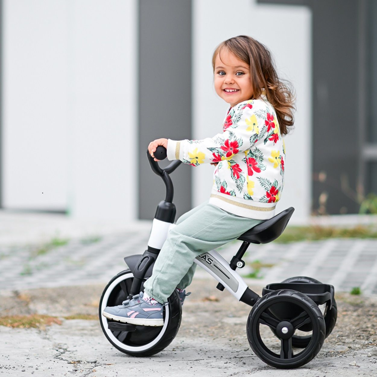 Una niña sonríe en triciclo. Lleva una chaqueta floral y pantalones verdes.