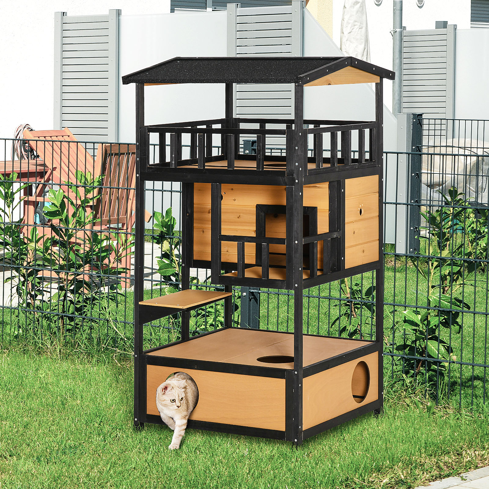 Ein mehrstöckiges Katzenhaus aus Holz, aus dem eine Katze kommt. Grünes Gras und Zaun sind zu sehen.