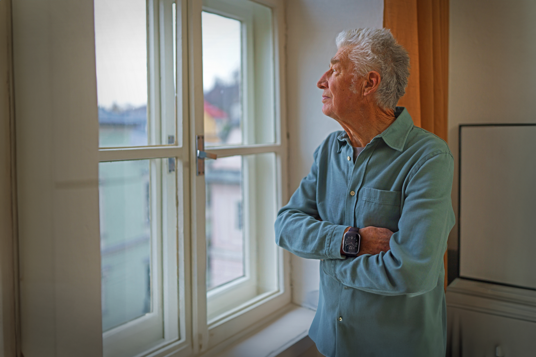 Ein älterer Mann schaut aus dem Fenster, Hände gekreuzt. Natürliches Licht und häusliche Umgebung.