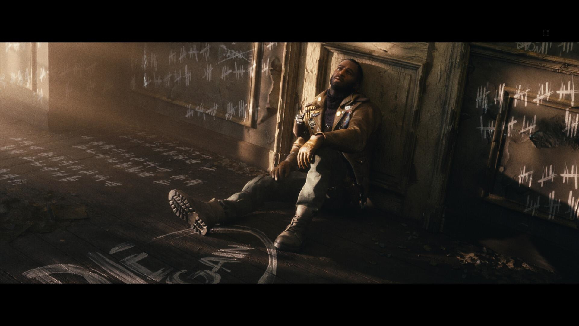 Mężczyzna siedzi na drewnianej podłodze pod ścianą pokrytą znakami. Światło pada na podłogę.