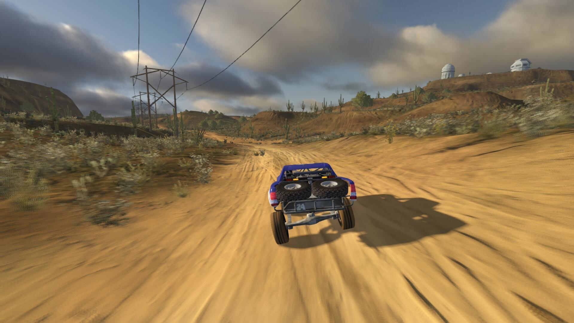 Ein blauer Truck fährt auf einer sandigen Straße mit Wüstenlandschaft und Stromleitungen.