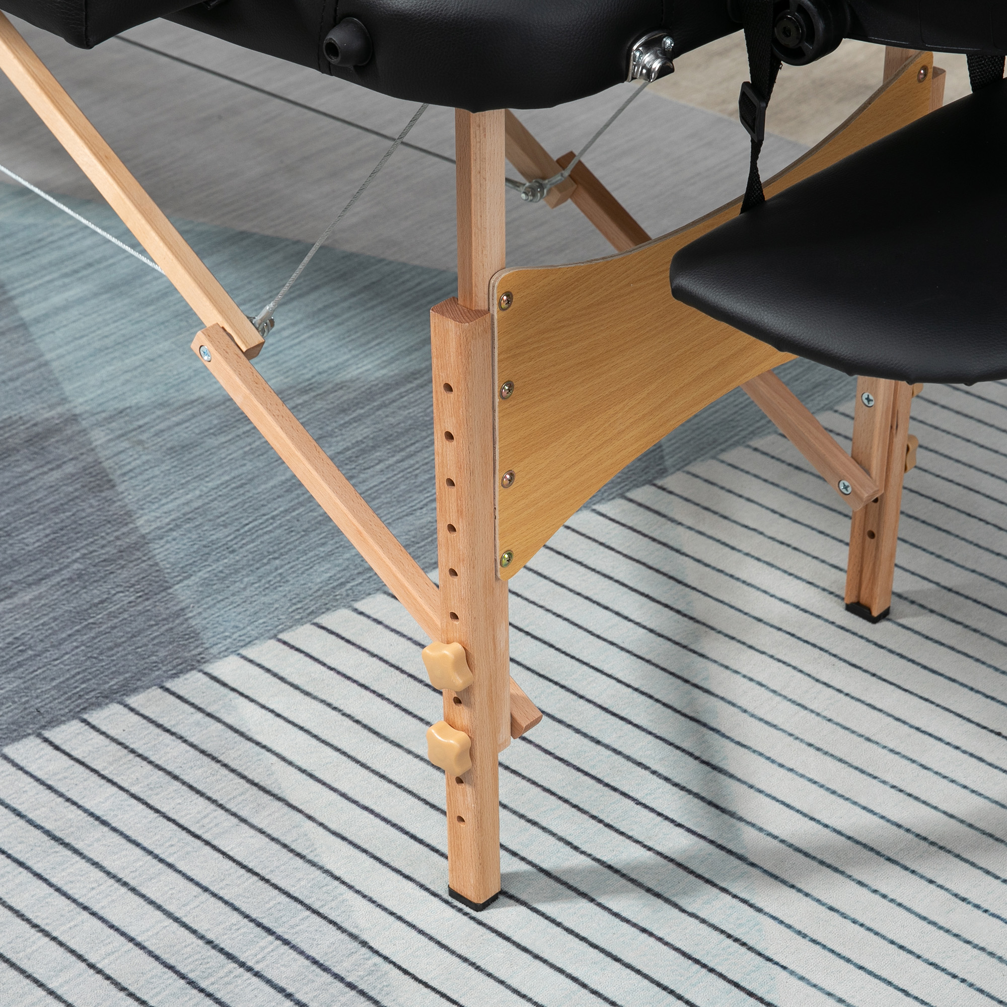Detalle de mesa de masaje: cojín negro, estructura de madera y alfombra rayada.