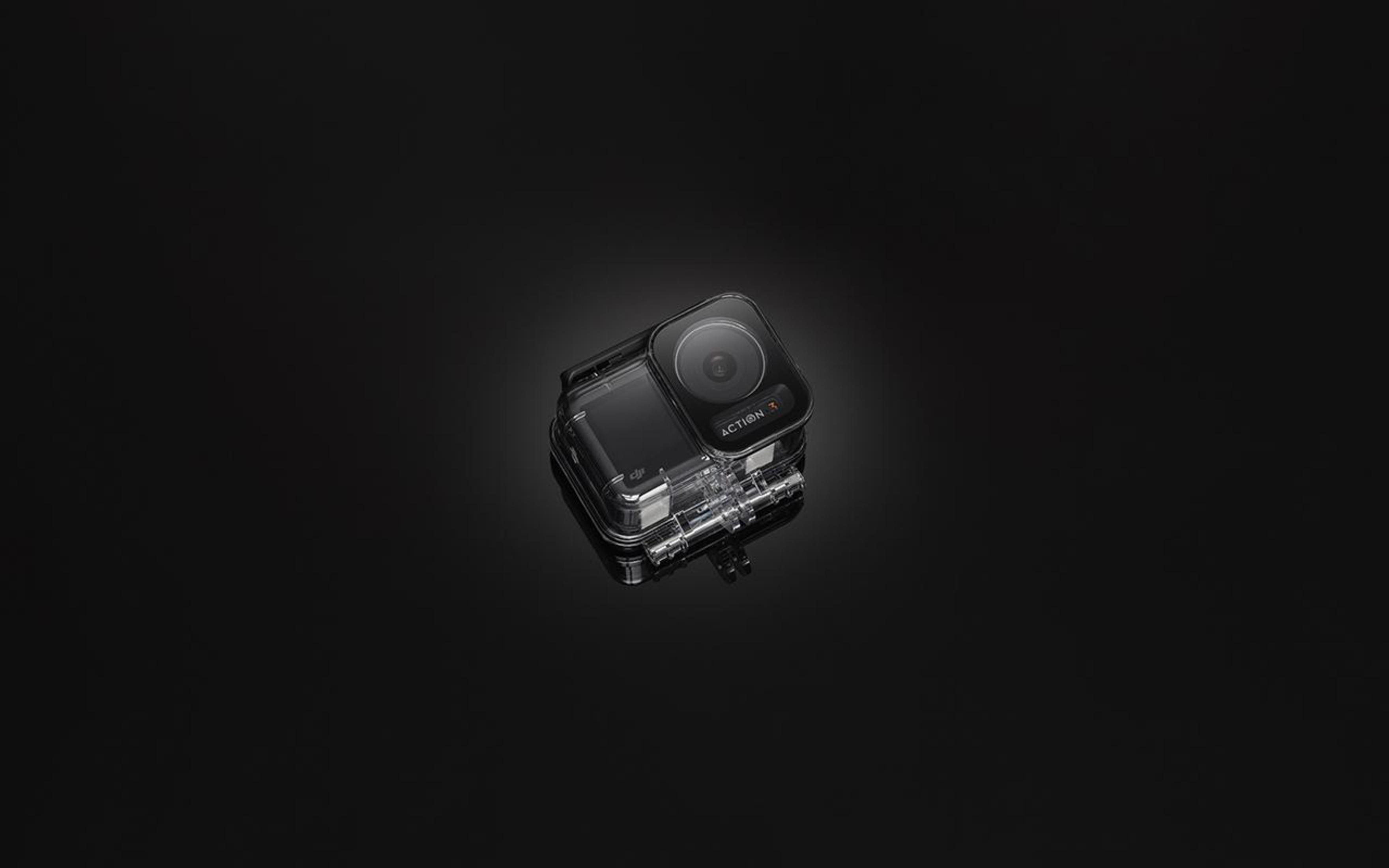 Mała przezroczysta obudowa kamery z obiektywem, na ciemnej powierzchni. Jest skierowana w stronę widza.