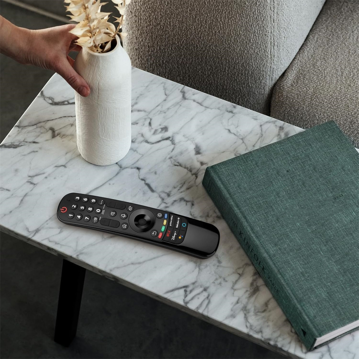 Fernbedienung auf einem Tisch, mit einem Buch und einer Vase mit Blumen.