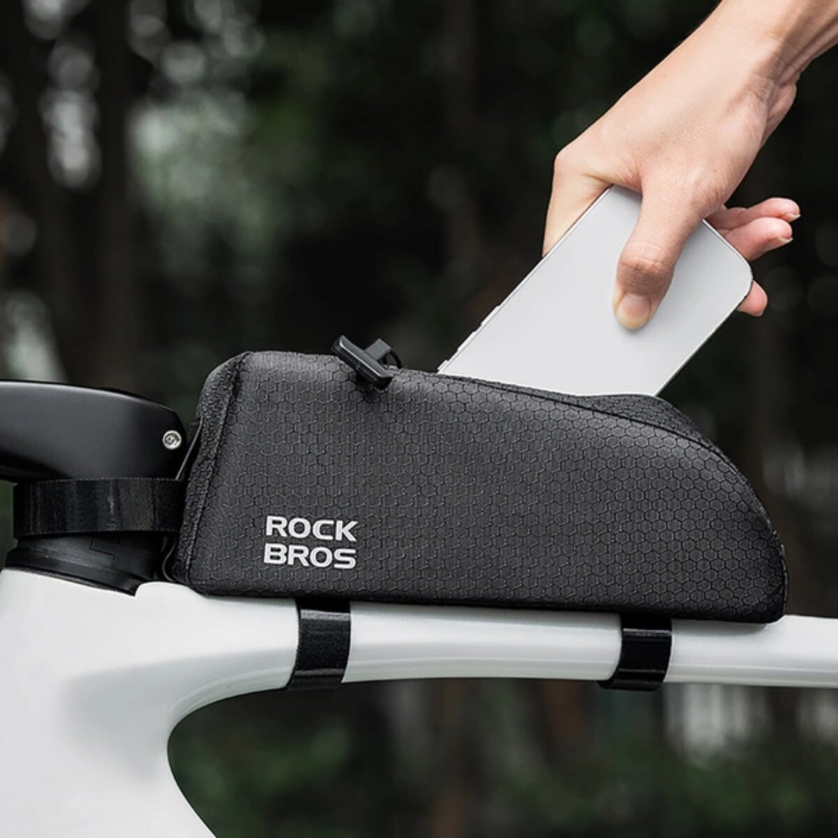 Hand legt ein Telefon in eine schwarze Rahmentasche mit ROCK BROS Logo auf einem weißen Fahrradrahmen.