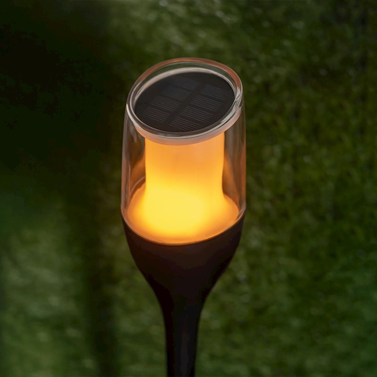 Une lampe solaire de jardin avec une lueur en forme de flamme. Dessus en plastique transparent, base noire.