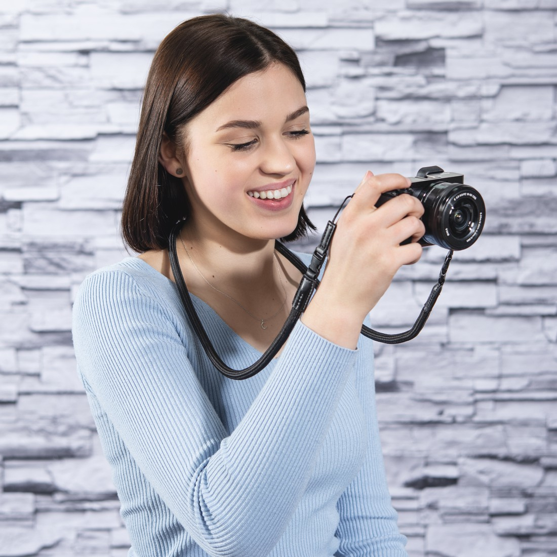 Młoda kobieta uśmiecha się trzymając czarny aparat z paskiem na szyję, stojąc przed ceglaną ścianą.