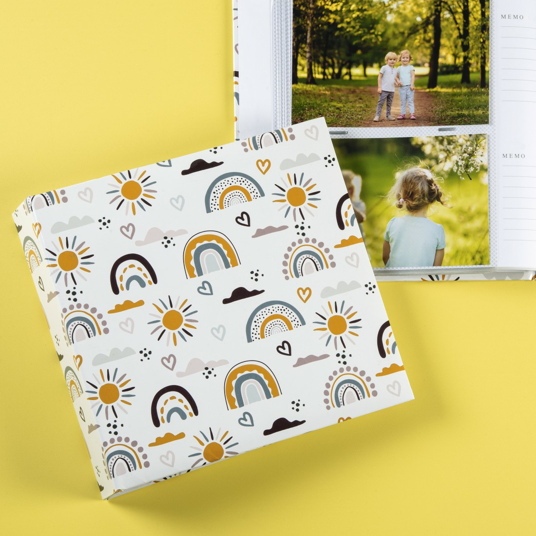 Otwarty album ze zdjęciami na żółtej powierzchni, z wzorzystą okładką przedstawiającą tęcze i słońca.