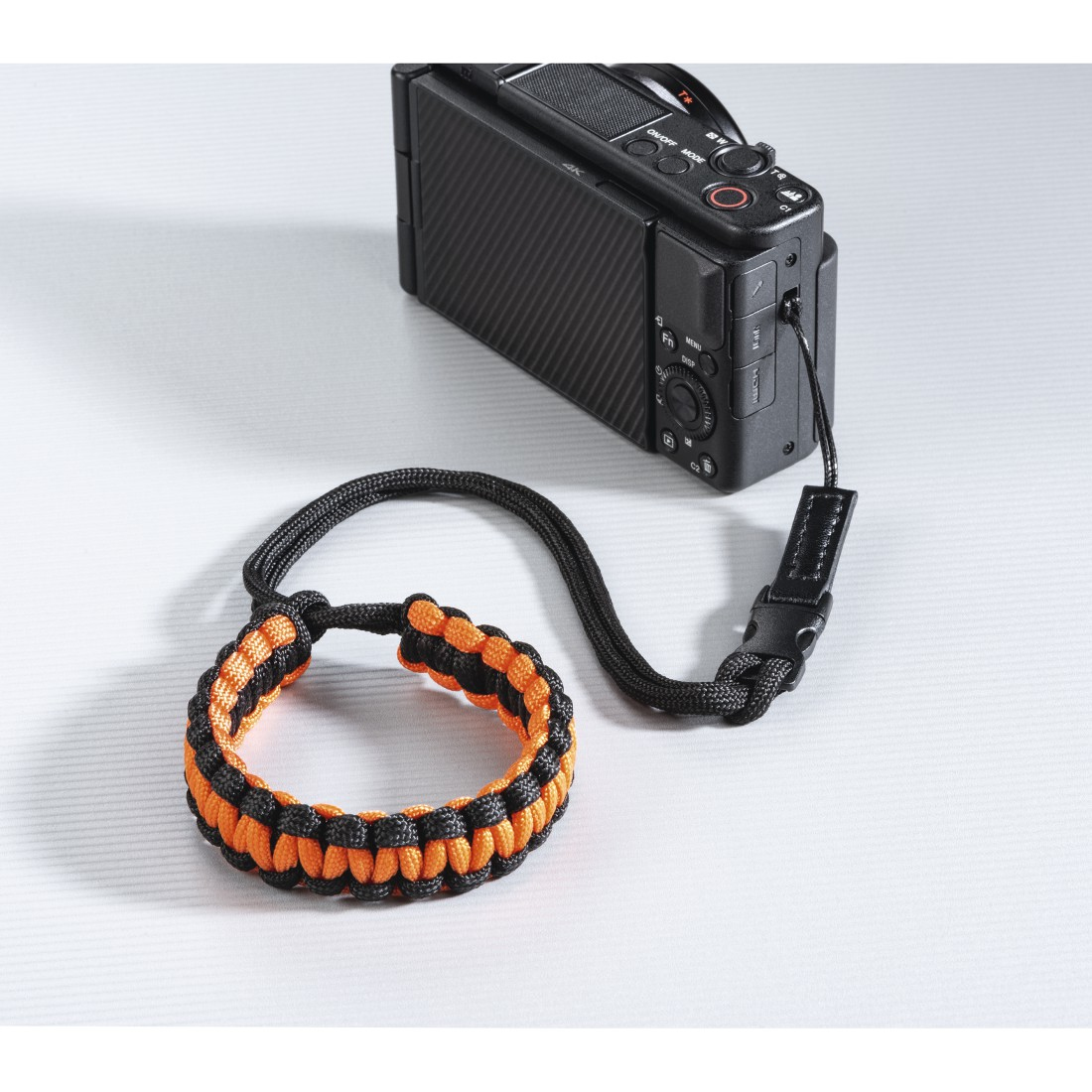Une dragonne noire et orange fixée à un appareil photo. La sangle est tissée et l'appareil photo est noir.