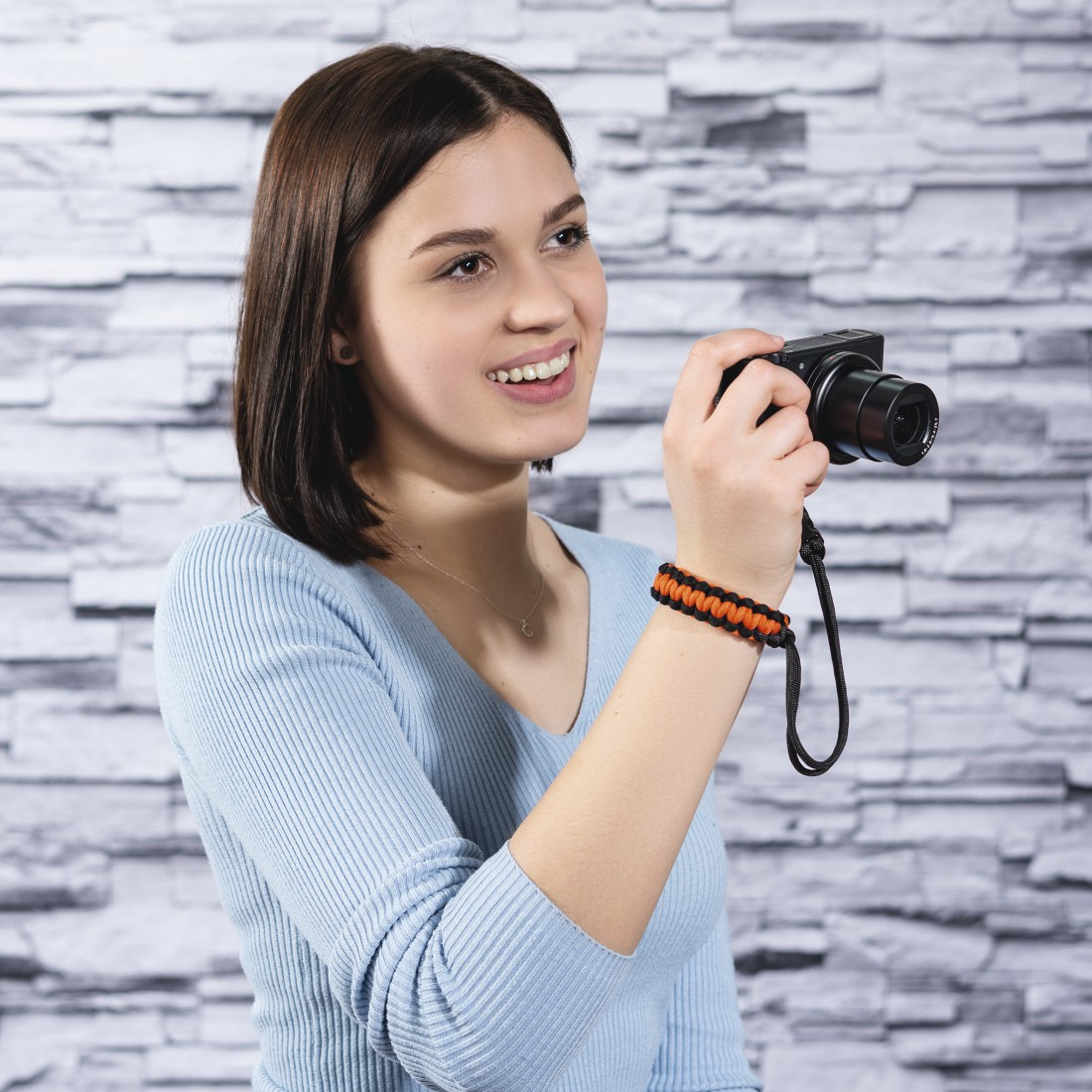 Une femme avec une dragonne d'appareil photo sourit. Elle porte une chemise bleue et tient un appareil photo.