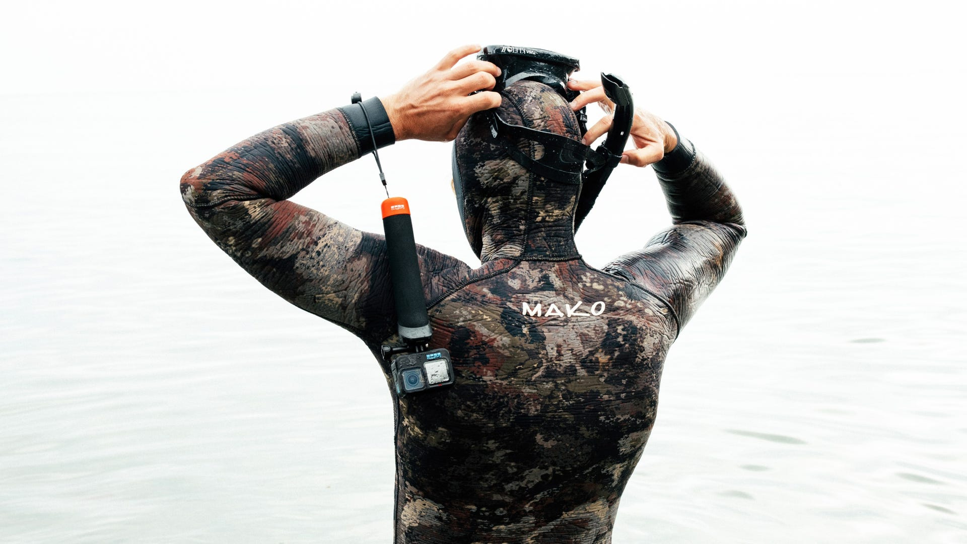 Osoba w kombinezonie nurkowym przygotowuje się do nurkowania. Ma maskę i trzyma kamerę GoPro z uchwytem.