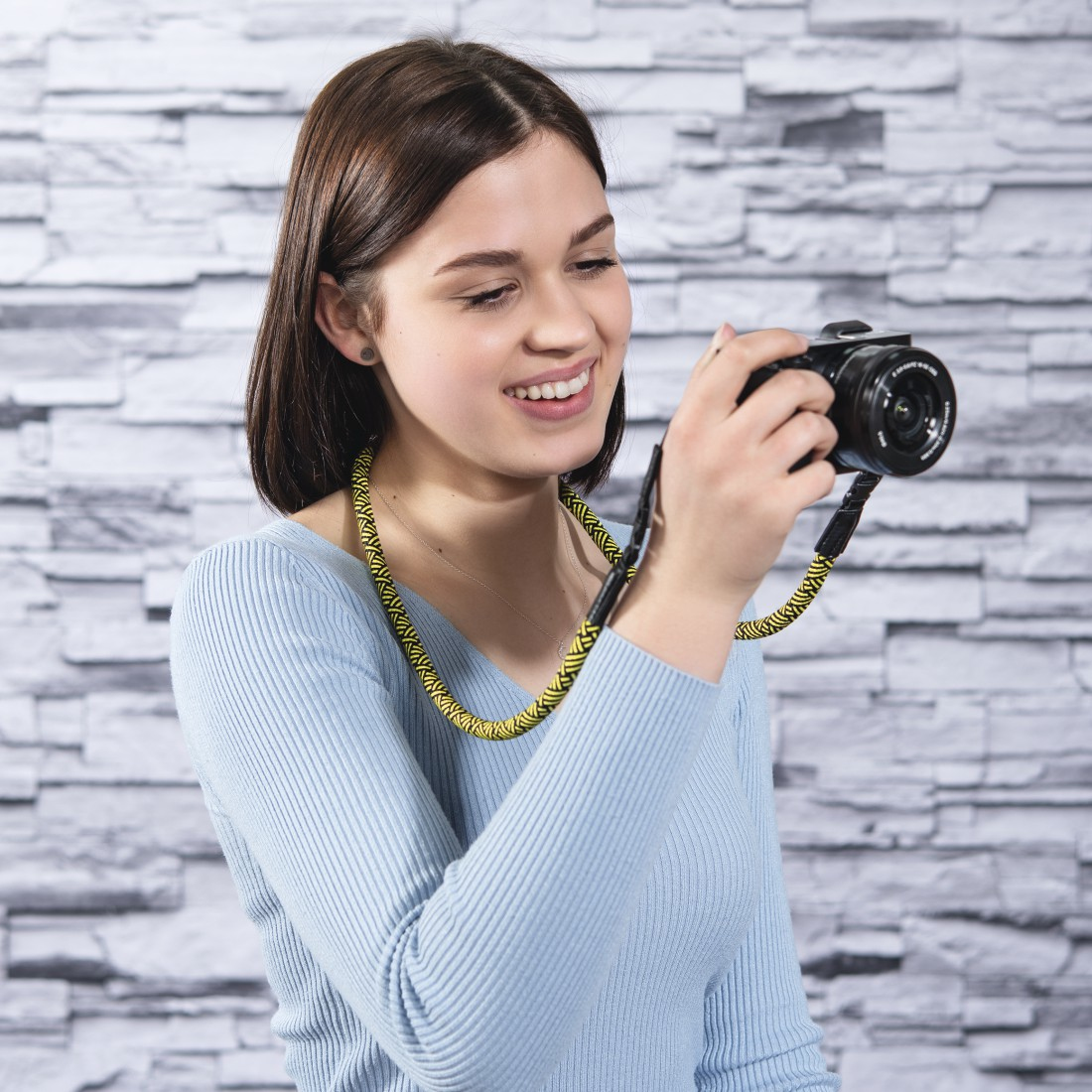 Uśmiechnięta kobieta trzyma aparat z żółto-czarnym paskiem, na tle ceglanej ściany.