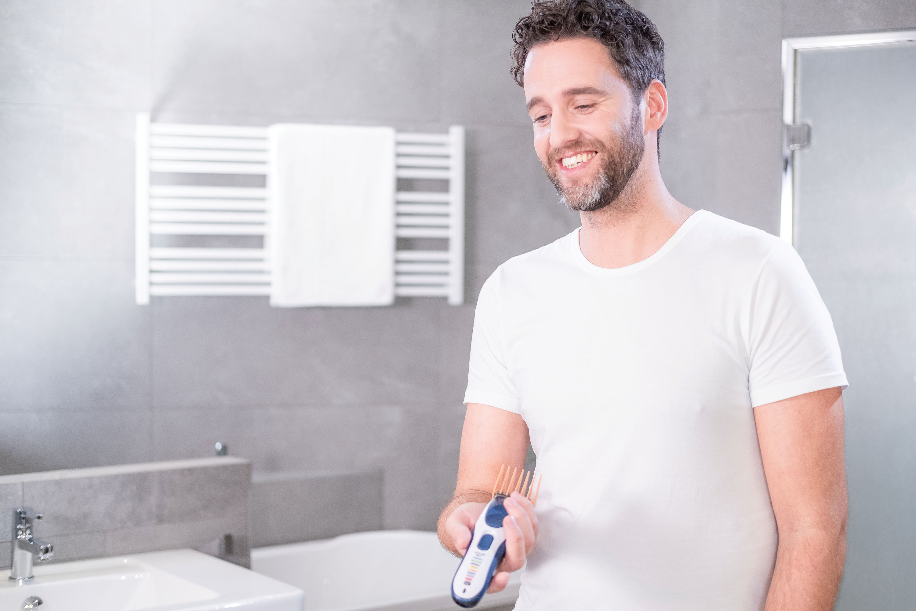 Hombre con camisa blanca sonríe, sosteniendo cortadora de pelo en un baño.