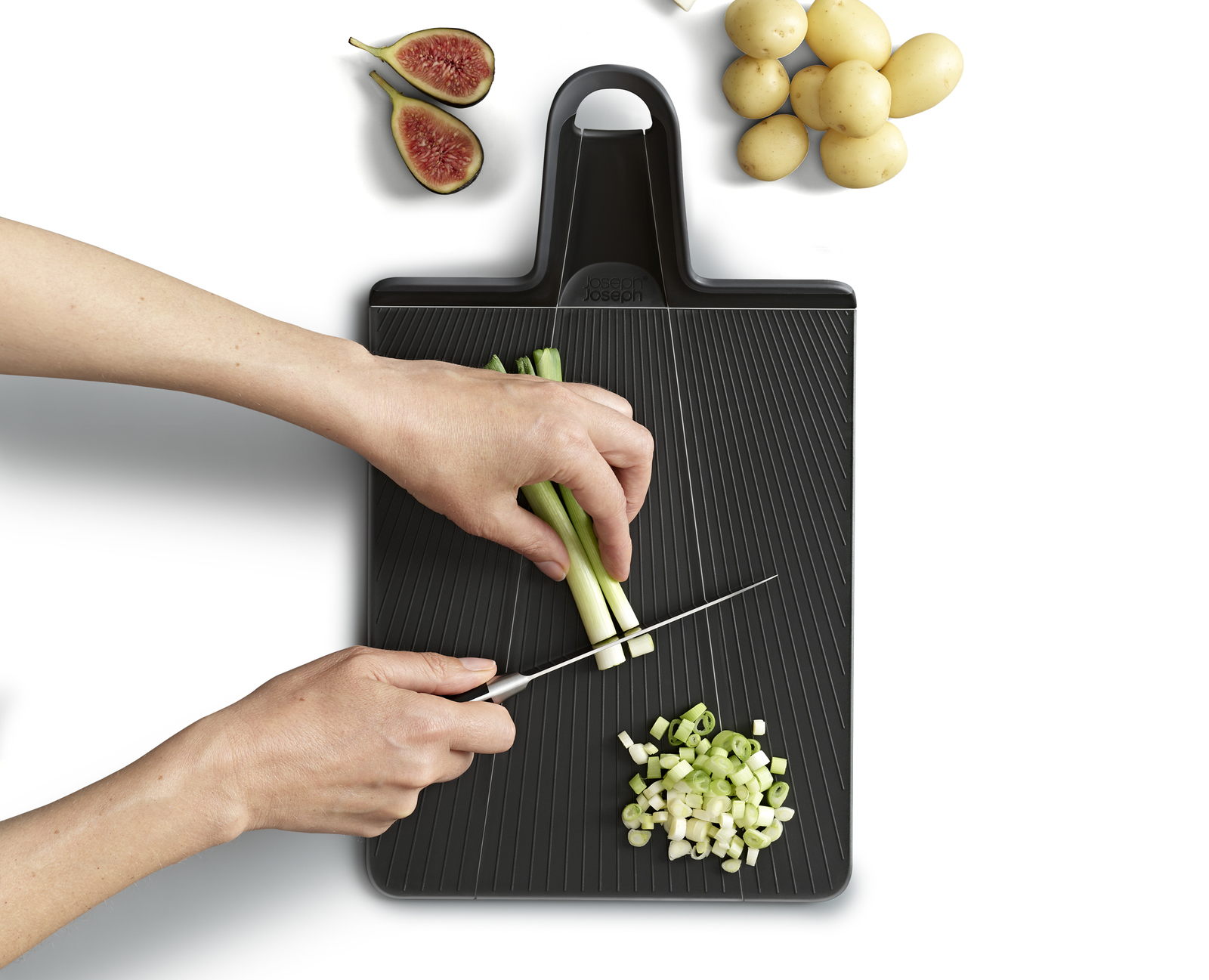 La imagen muestra a una persona cortando cebolletas en una tabla de cortar negra, con higos y patatas cerca.