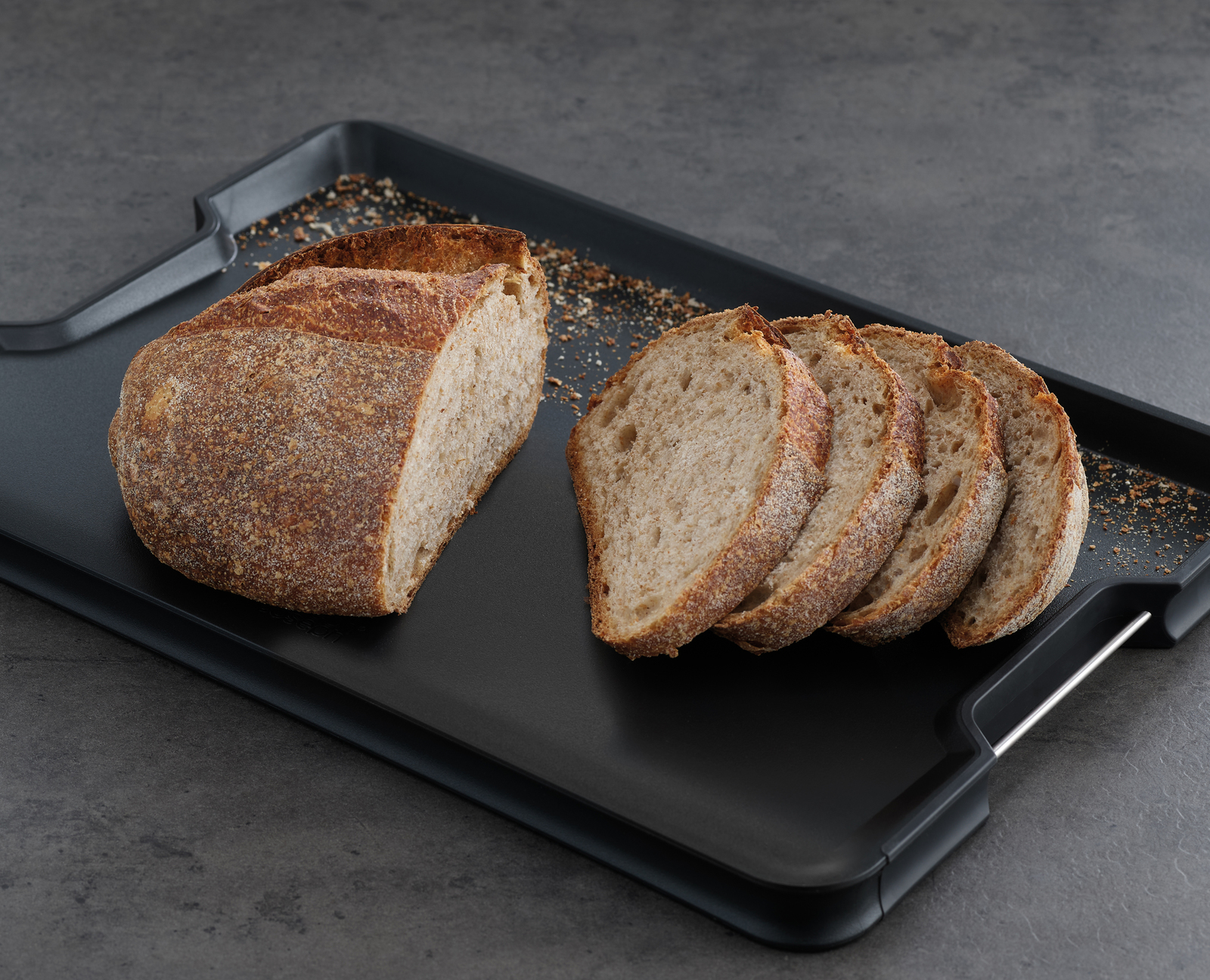 Brotlaib und Scheiben auf schwarzem Tablett. Das Brot hat ein rustikales Aussehen.