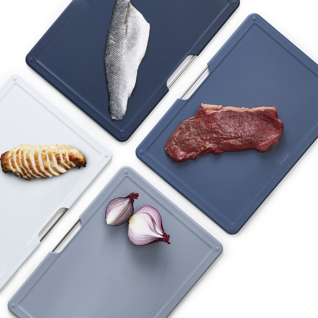 Cuatro tablas de cortar con alimentos. Tabla blanca con pollo, azul con pescado, gris con cebolla y azul con bistec.
