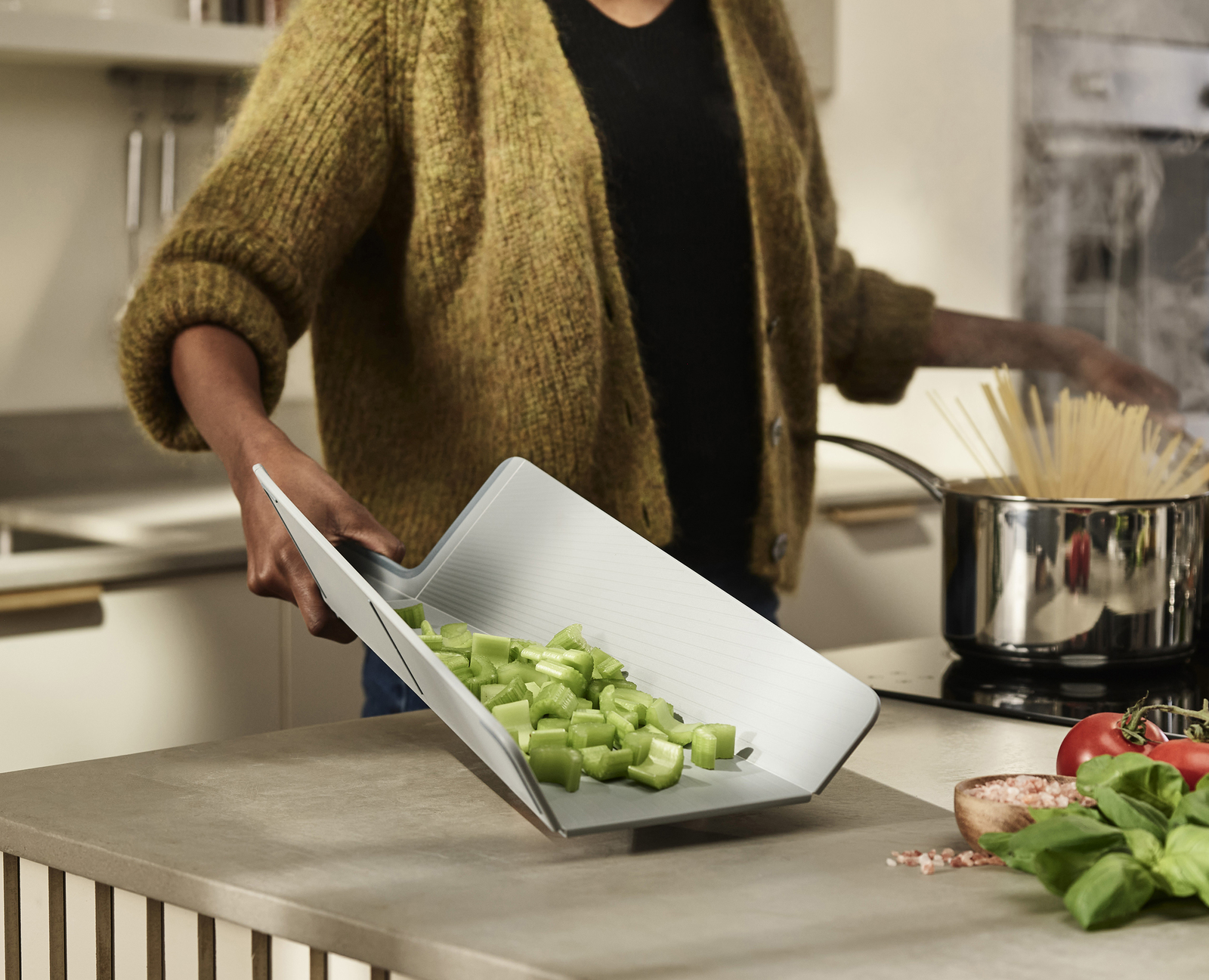 Una persona sostiene una tabla de cortar gris con verduras verdes picadas. Una olla con pasta y otros ingredientes están en el fondo.