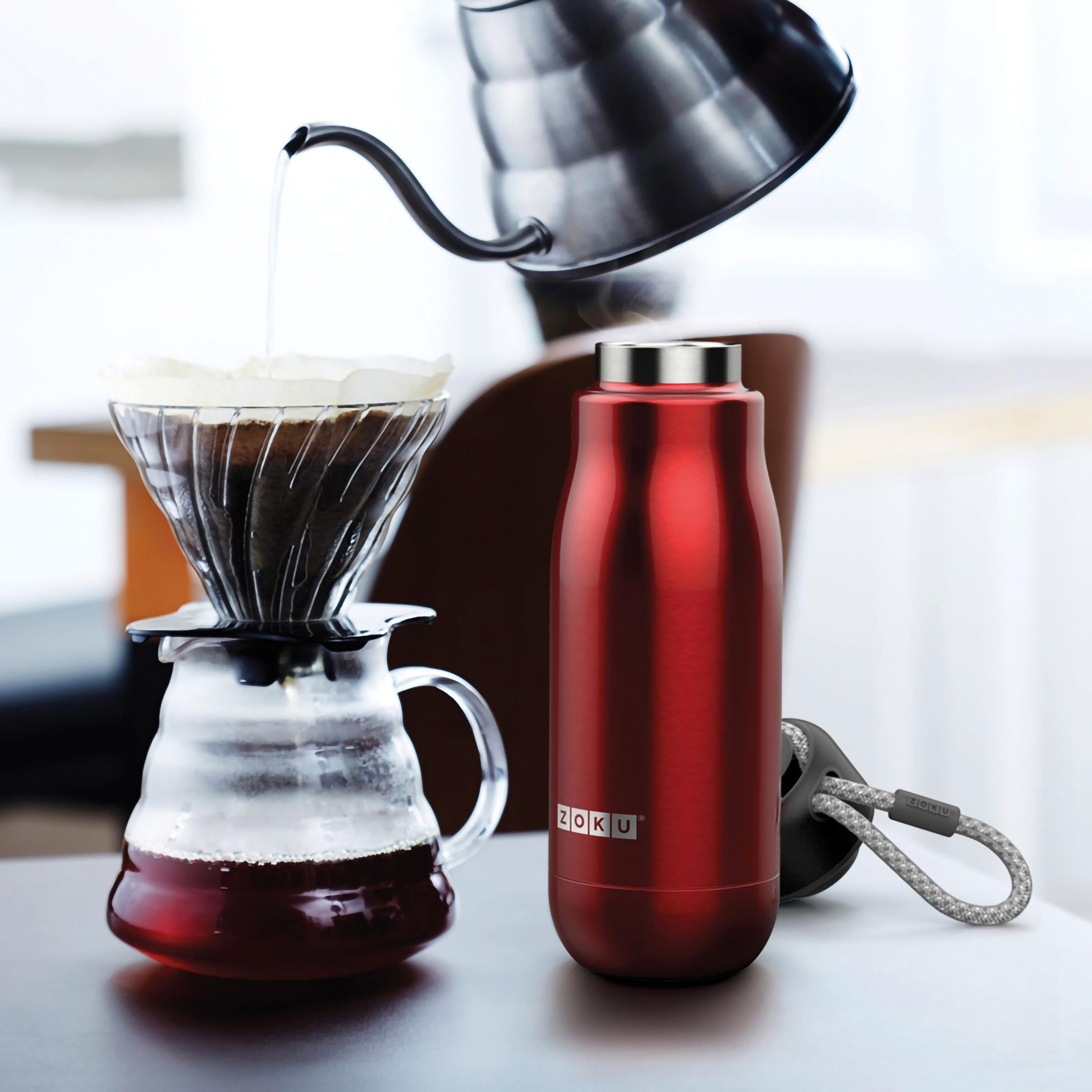Rote Wasserflasche, Kaffee wird in ein Glas gegossen. Eine ZOKU-Wasserflasche und eine Kaffeemaschine sind abgebildet.