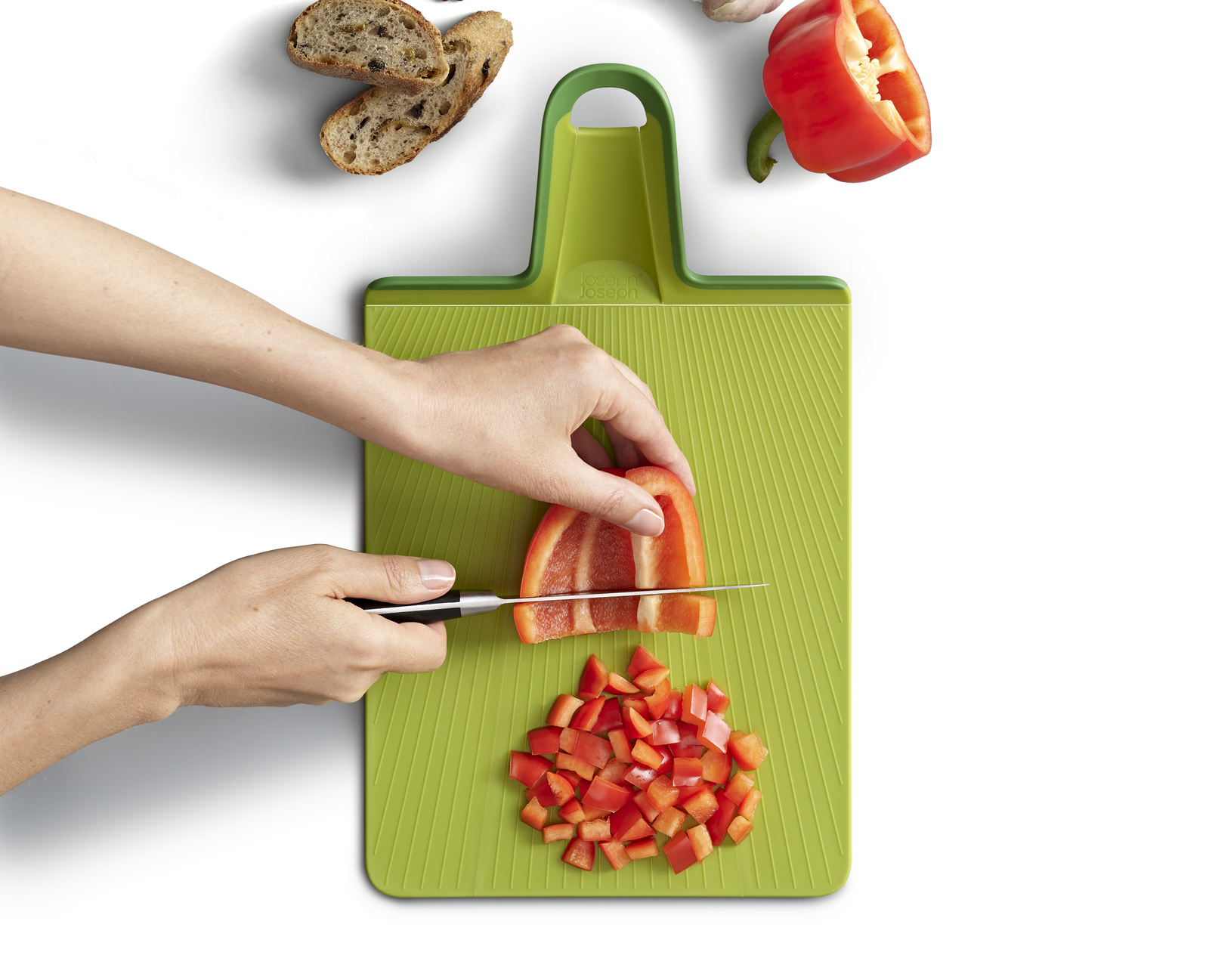 Eine Hand schneidet eine rote Paprika auf einem grünen Schneidebrett mit anderen Paprikastücken und Brot.