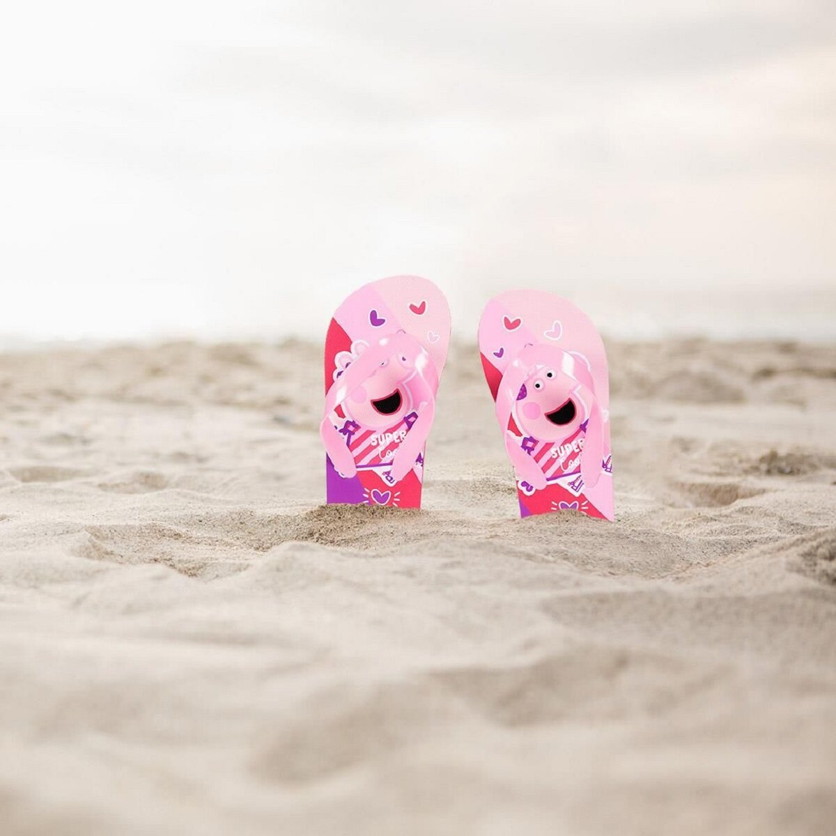 Zwei rosa Peppa Pig Flip-Flops an einem Sandstrand, mit verschwommenem Hintergrund.
