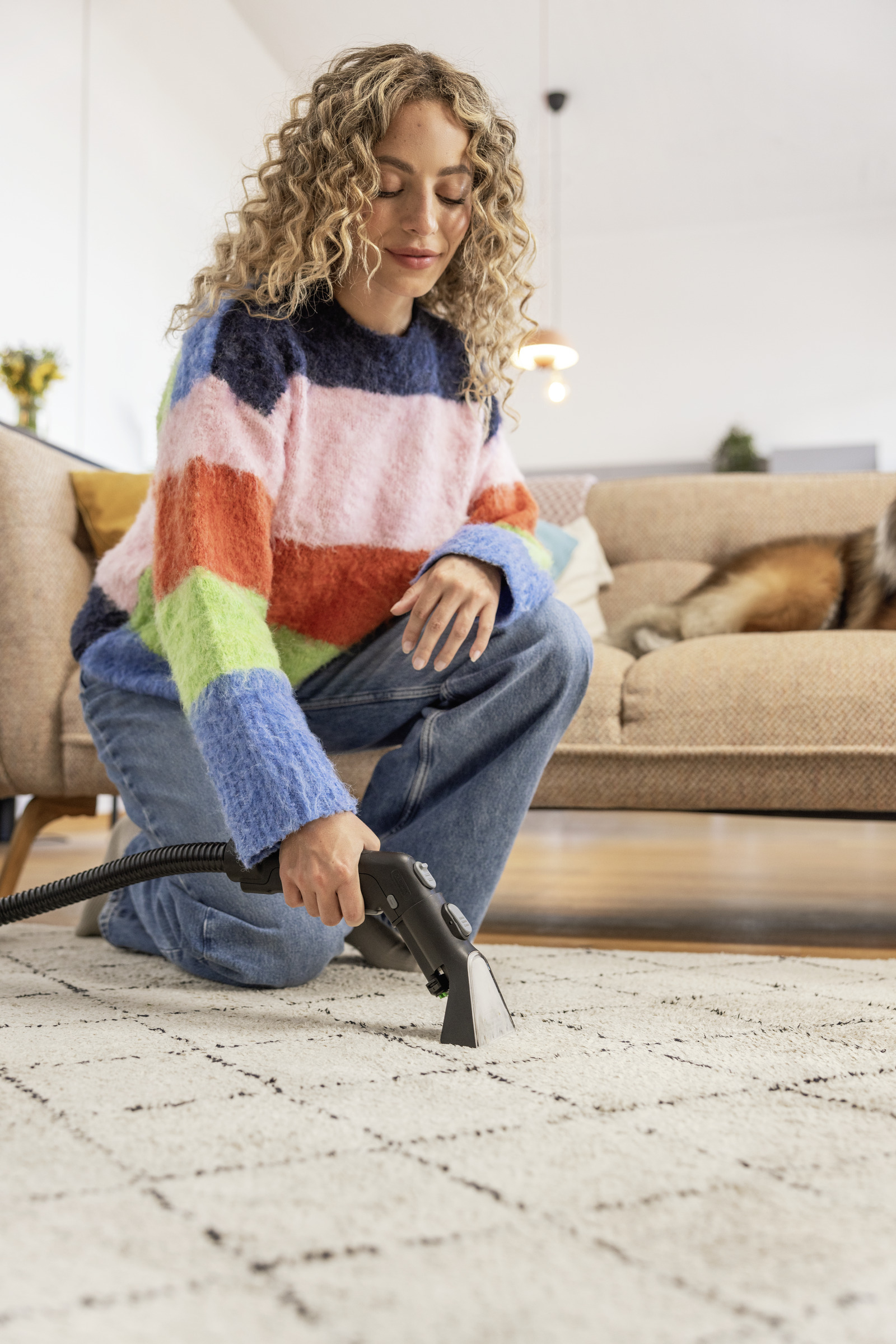 Kobieta czyści dywan parownicą. Klęczy na dywanie, w pobliżu sofy.