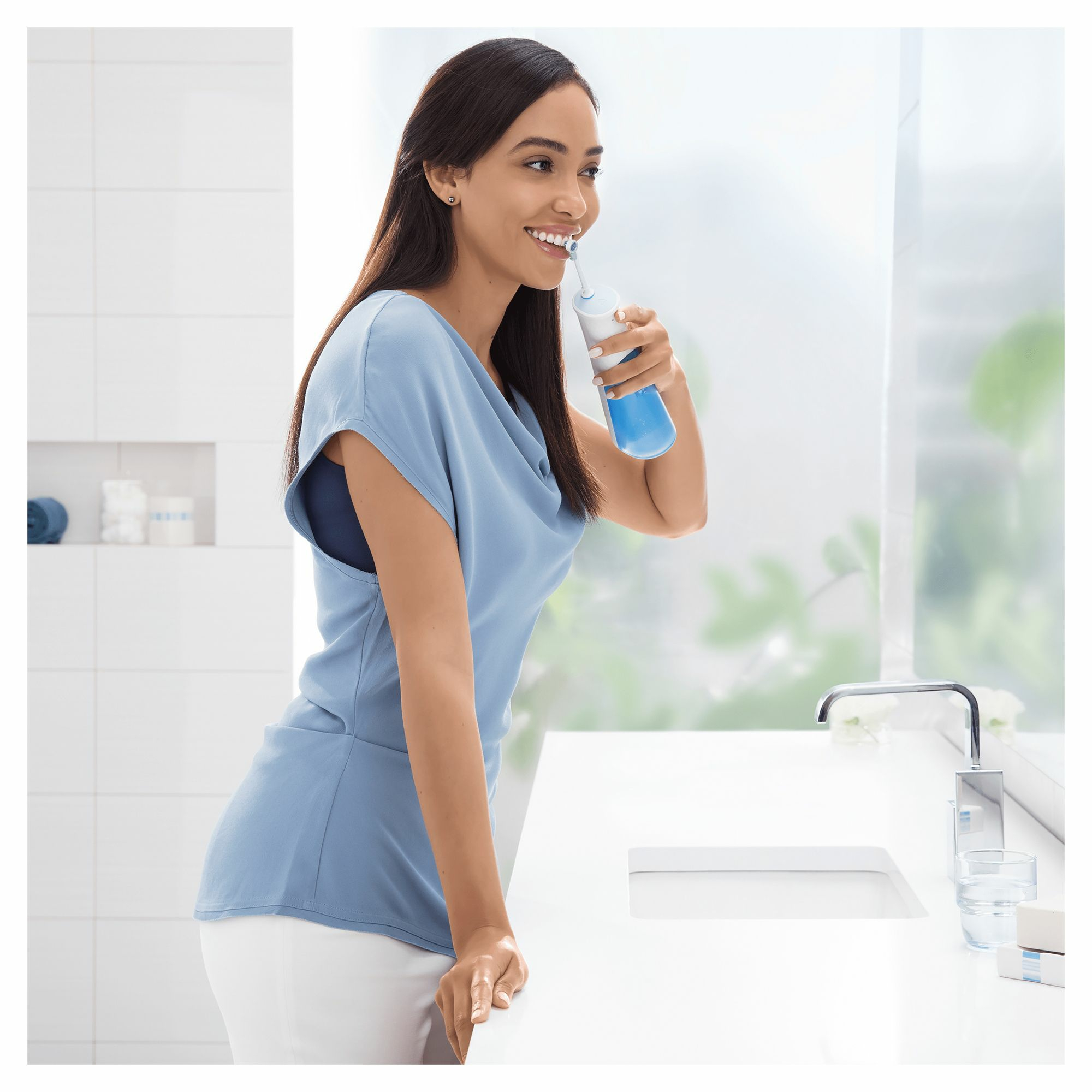 Una mujer usa un irrigador bucal azul y blanco en un baño, cerca de una ventana y un lavabo blanco.