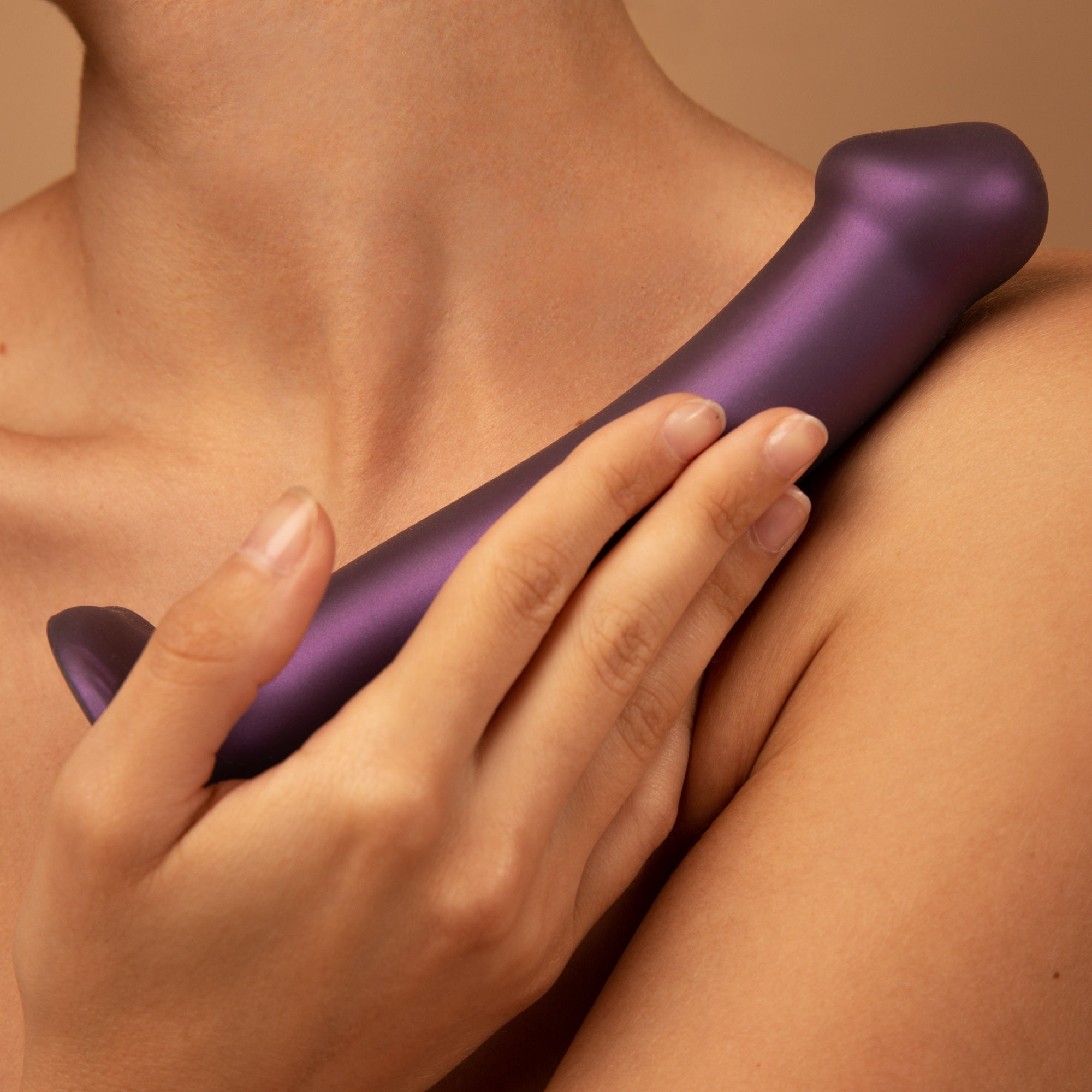 Ręka trzymająca fioletową zabawkę erotyczną na ramieniu. Tło jest beżowe.