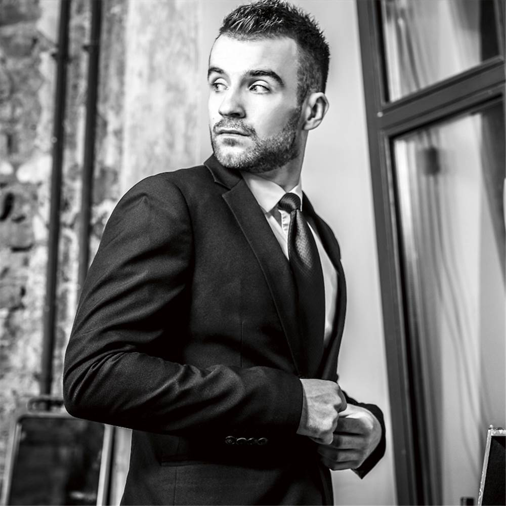 Un uomo in abito nero, guarda sopra la spalla. Muro di mattoni e finestra sullo sfondo.