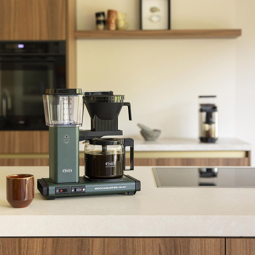 Scena kuchenna z ekspresem do kawy, szklankami i kwiatami na blacie w pobliżu okna.