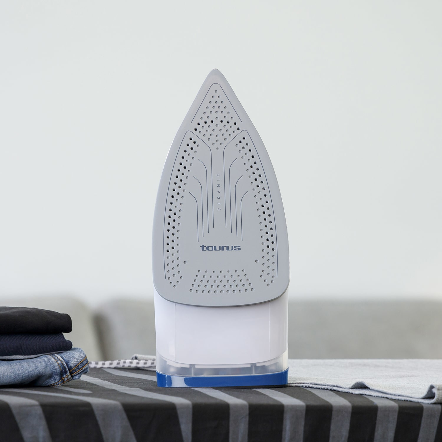 Szare żelazko na paskowanej powierzchni. Stos ubrań i sofa w tle.