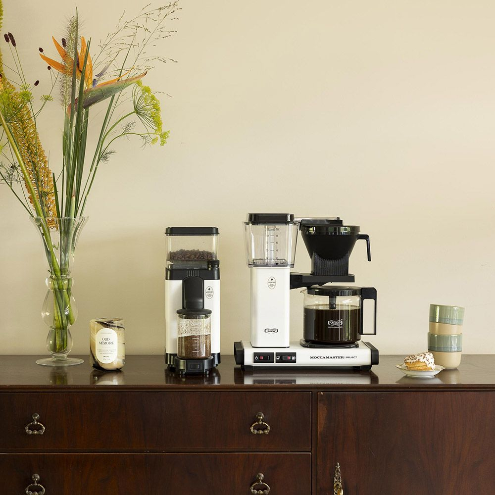 Szafka z ekspresem do kawy, kwiatami w wazonie, świecami i talerzem z pączkiem i filiżankami. Przytulne otoczenie.