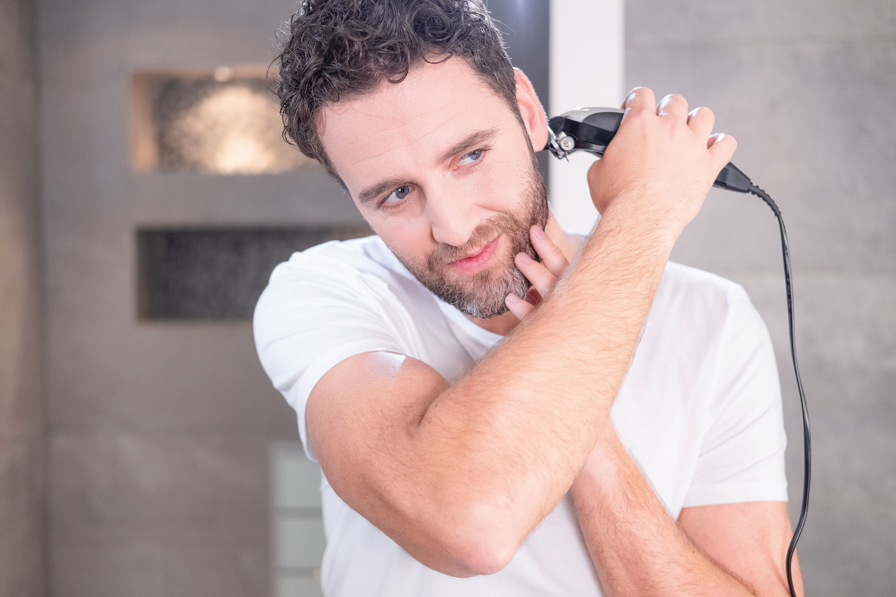 Mężczyzna przycina brodę czarnym trymerem przed lustrem, na jasnym tle.