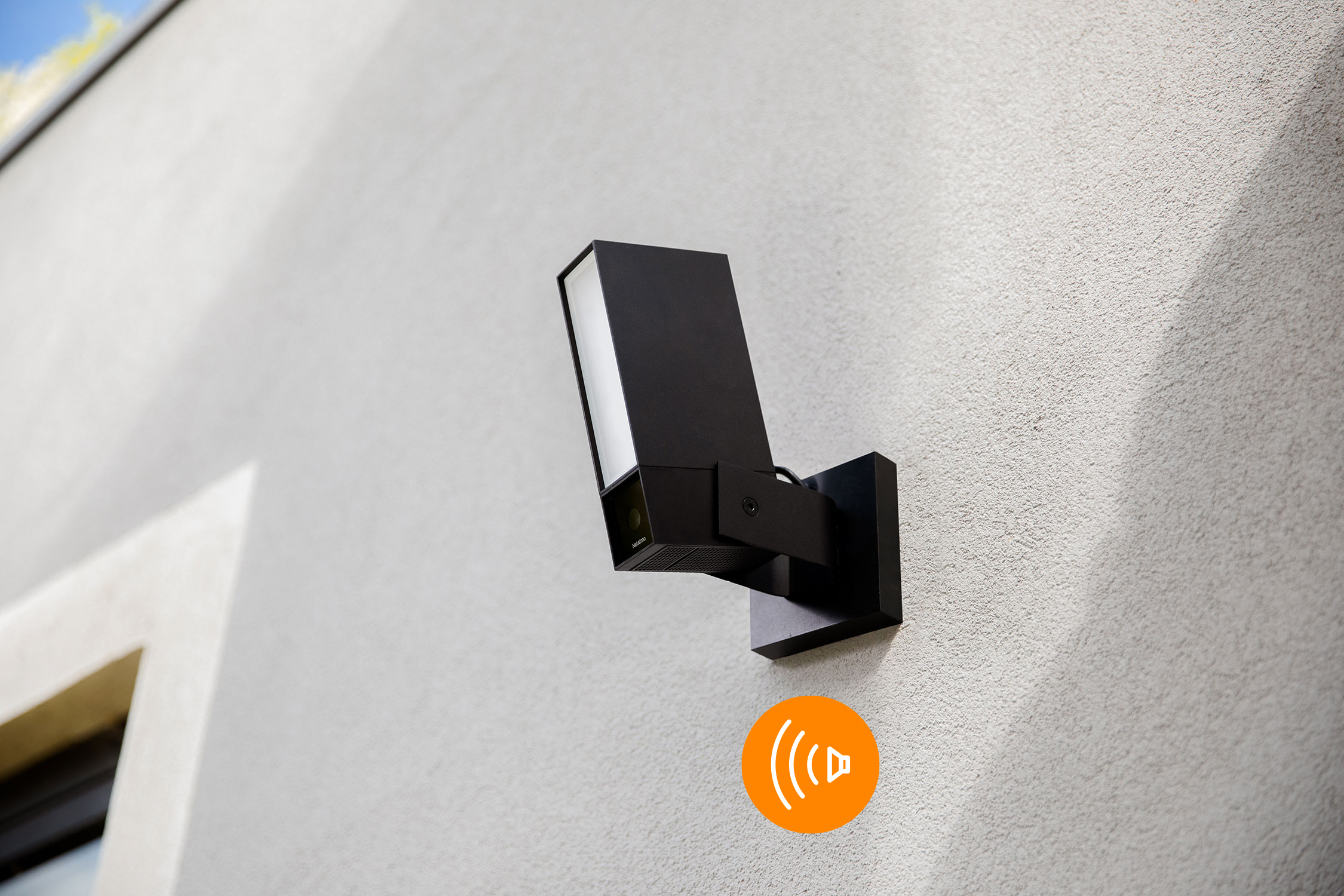 Una telecamera di sicurezza Netatmo nera montata su un muro grigio. C'è un'icona di altoparlante arancione.