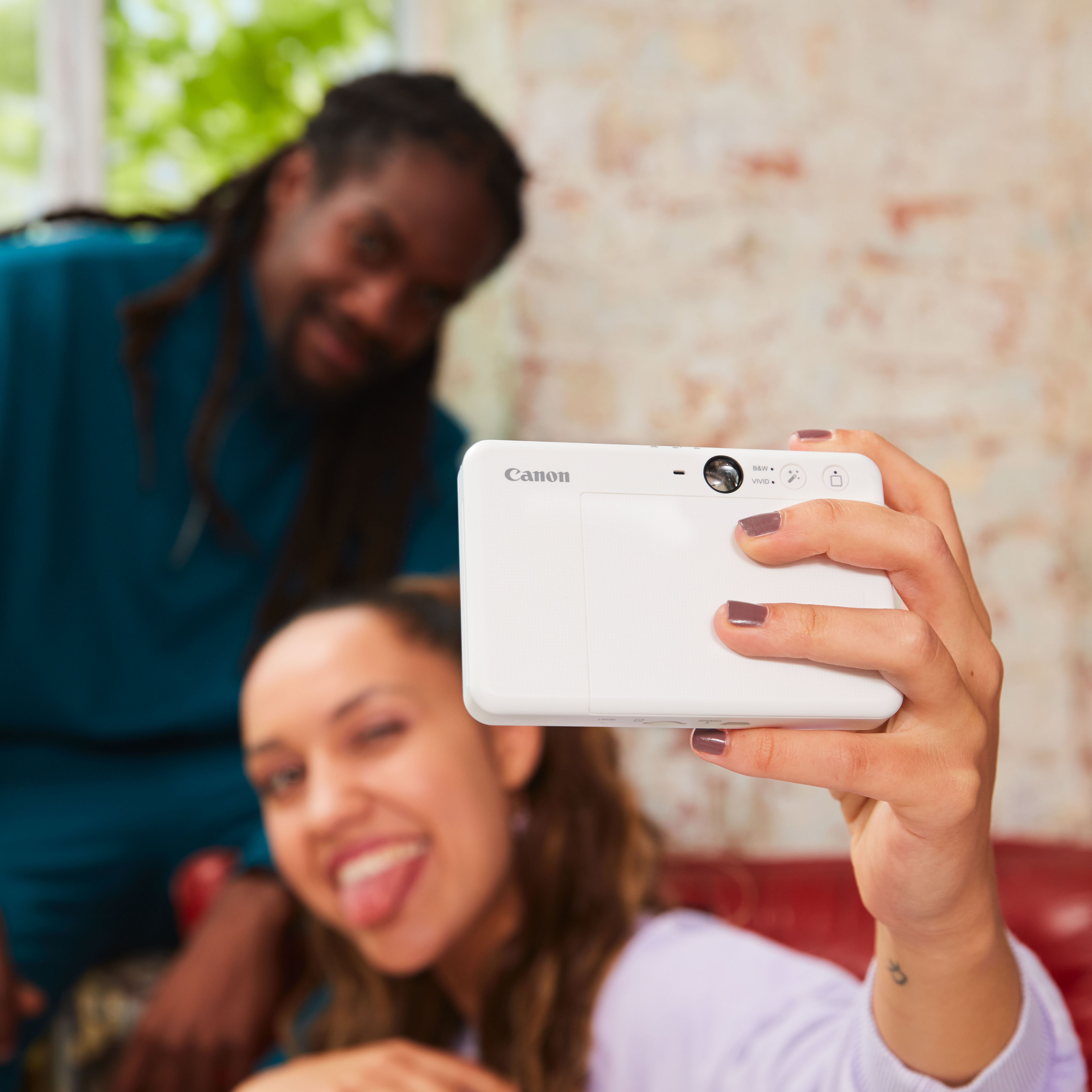 Kobieta robi selfie białą kamerą, w tle uśmiecha się mężczyzna.