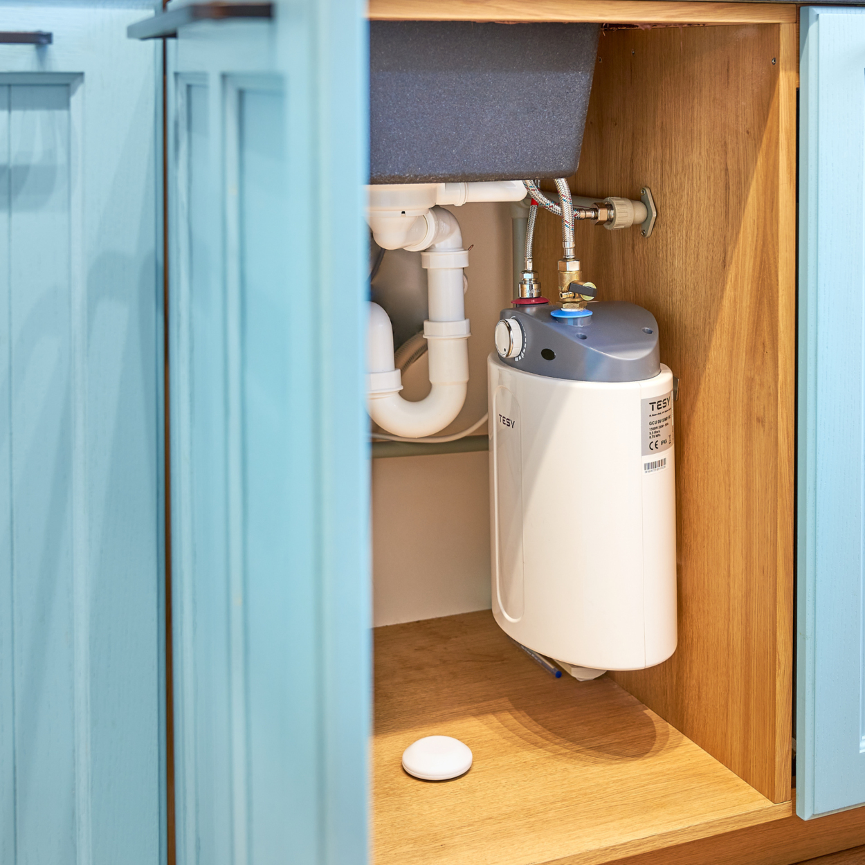 Ujęcie wnętrza szafki z podgrzewaczem wody i widoczną instalacją wodociągową, w otoczeniu kuchni.