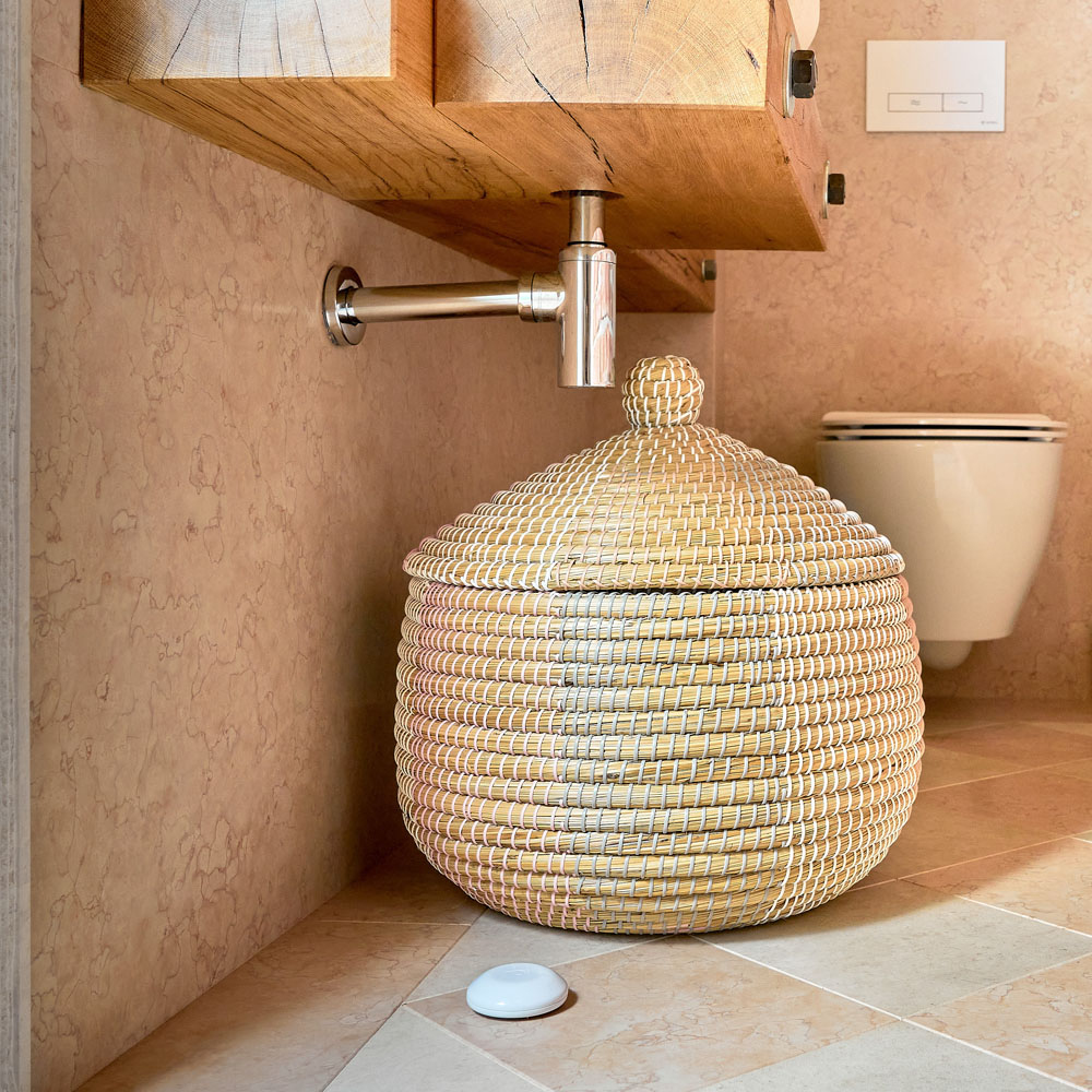 Tkany kosz z pokrywką stoi pod drewnianą toaletką. Mały biały przedmiot leży na podłodze.