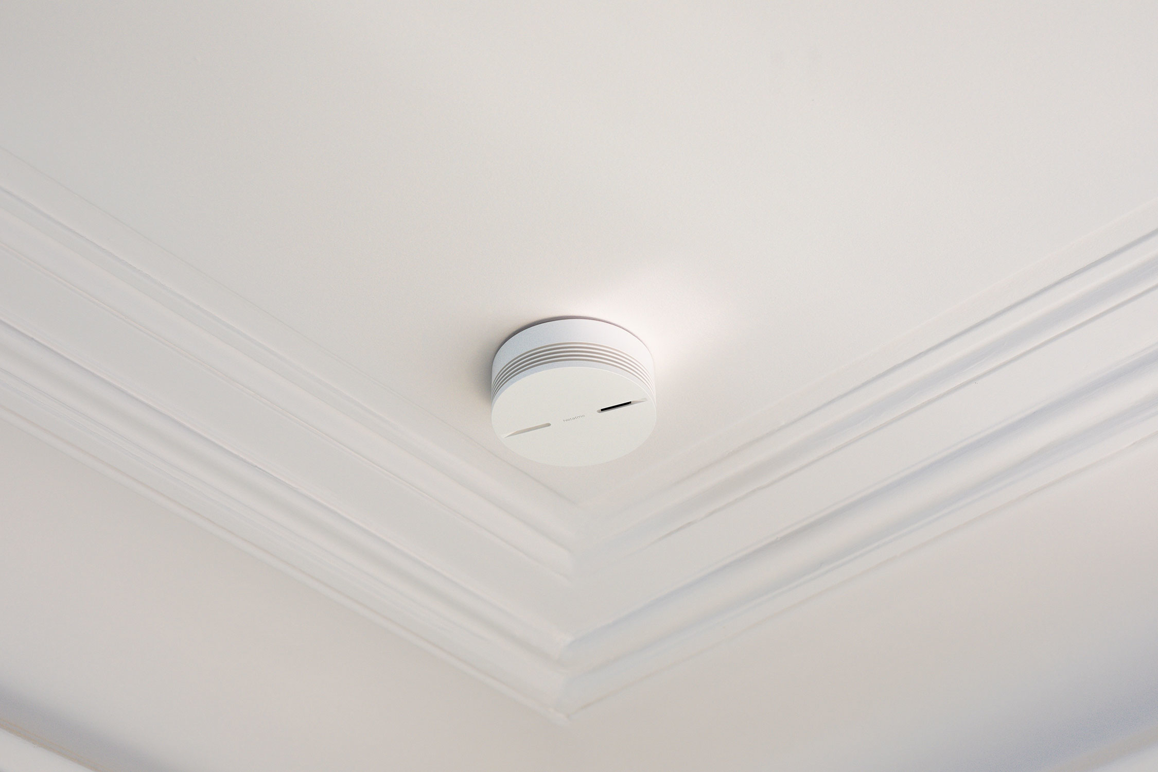 Witte rookmelder gemonteerd op een wit plafond. Het plafond heeft decoratieve lijsten.