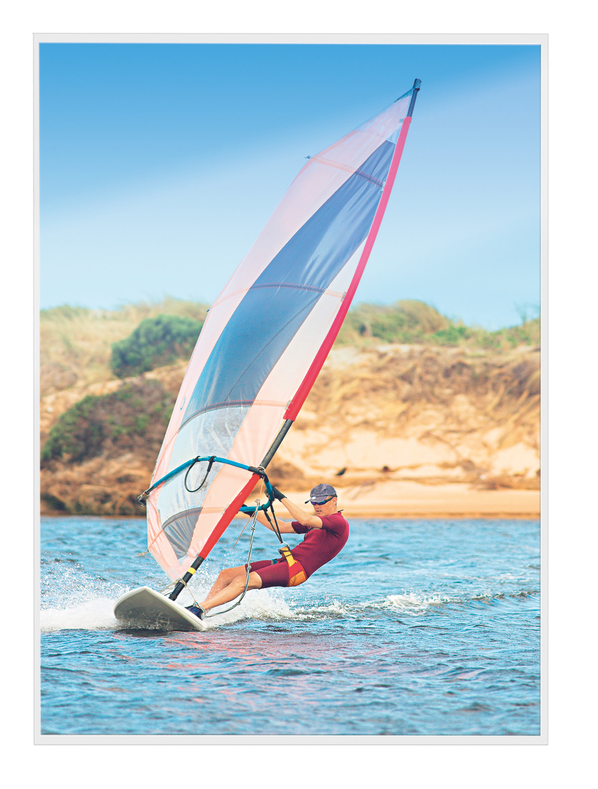 Eine Person beim Windsurfen auf blauem Wasser. Ein farbenfrohes Segel ist oben. Der Hintergrund hat Strand und Himmel.