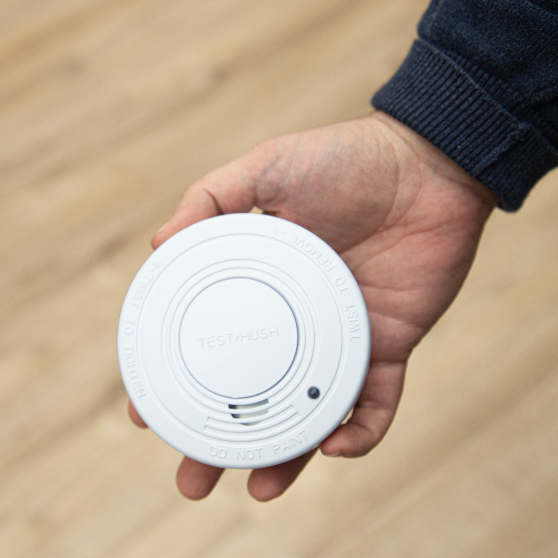 Ręka trzymająca biały czujnik dymu. Widoczne są słowa "TEST/HUSH".