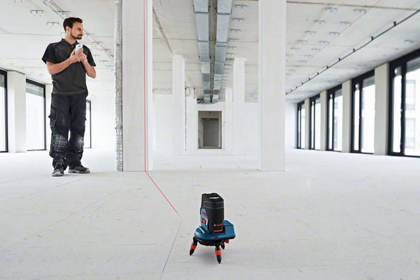 Mann verwendet Laser-Nivelliergerät in großem Raum. Der Laser projiziert eine rote Linie. Hintergrund hat weiße Wände, Decken und Säulen.