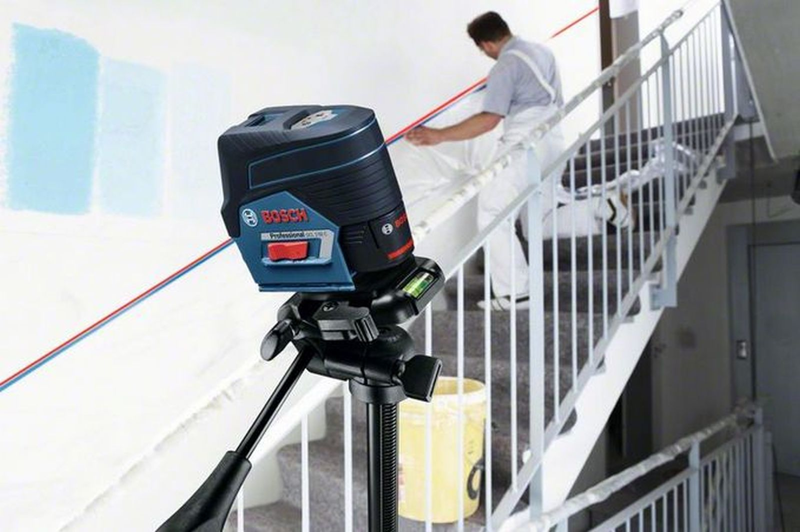 Ein Laser-Nivelliergerät auf einem Stativ projiziert eine rote Linie über Treppen. Ein Mann in Overalls bemalt die Wand.
