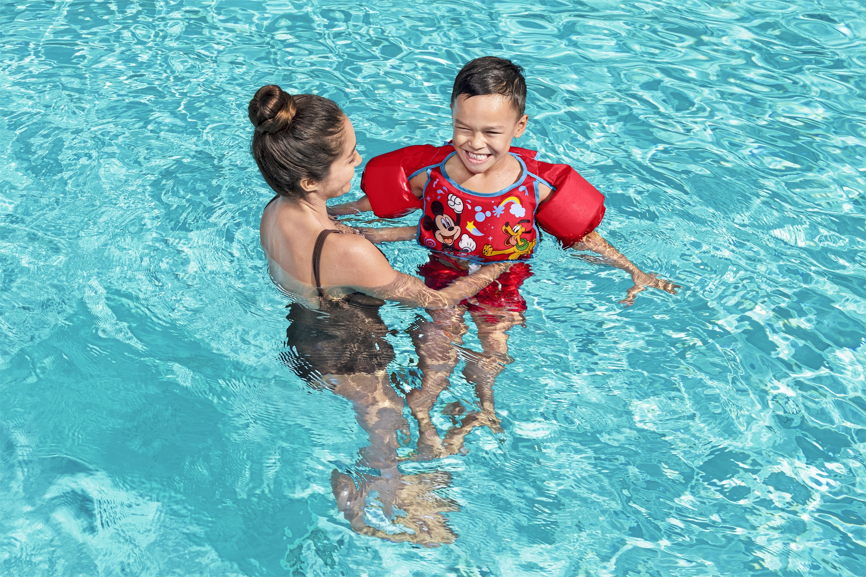 Kobieta i dziecko w basenie. Dziecko w czerwonych rękawkach, uśmiechnięte. Kobieta trzyma dziecko, oboje w niebieskiej wodzie, słoneczny dzień.