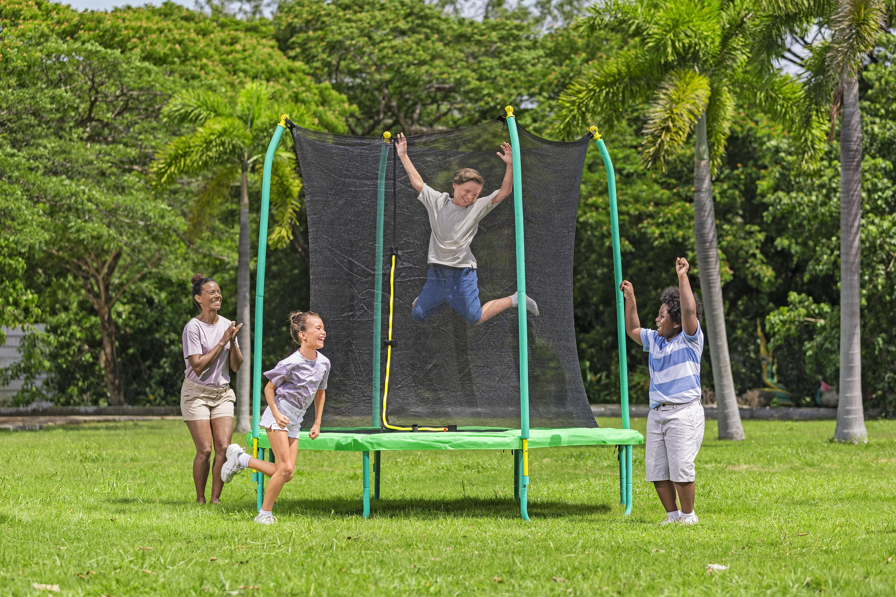 Cztery dzieci bawią się na trampolinie, jedno skacze. Zielona trawa, drzewa w tle, słoneczny dzień.