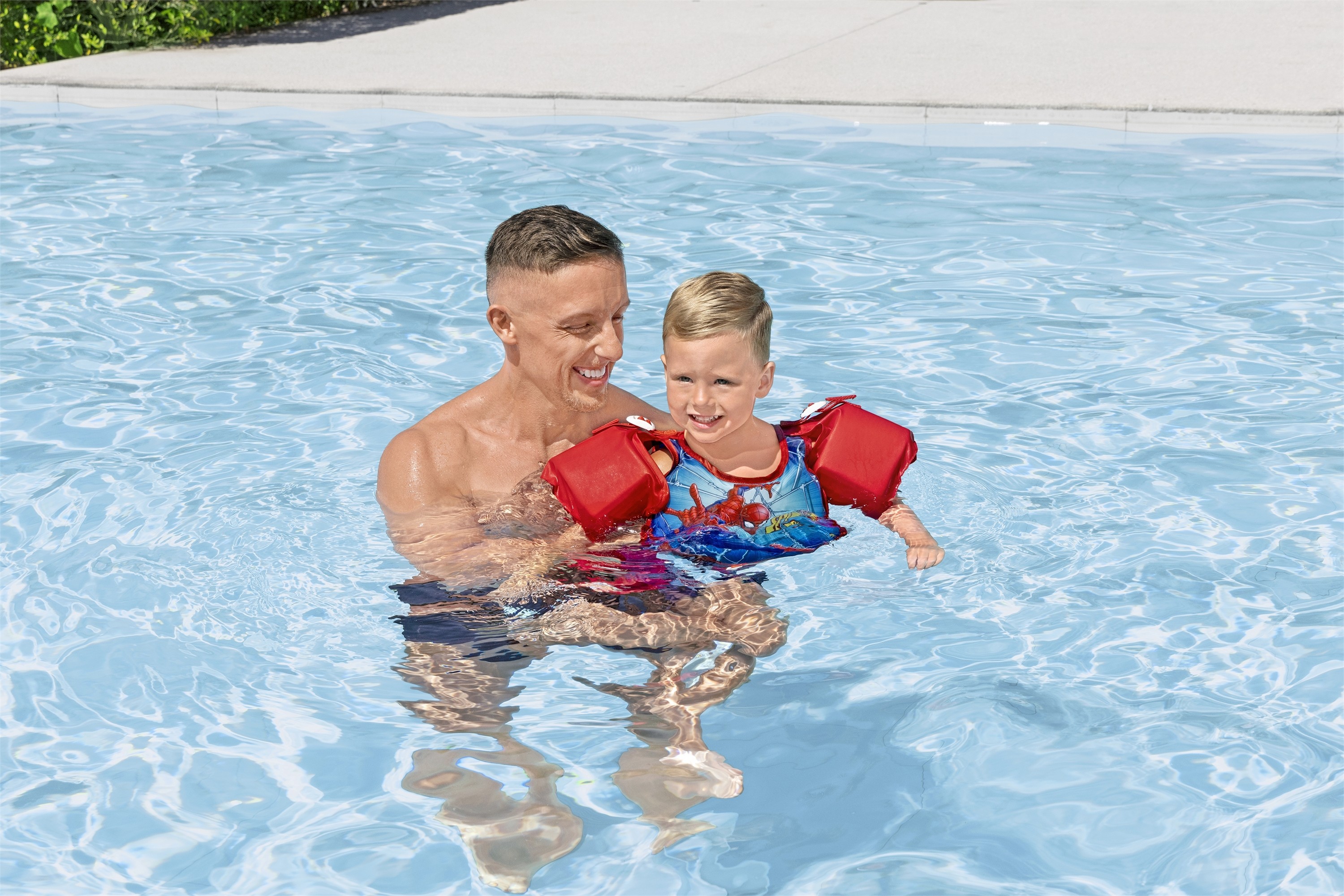 Uomo che tiene un bambino in acqua; entrambi sorridono; il bambino ha braccioli di Spider-Man.