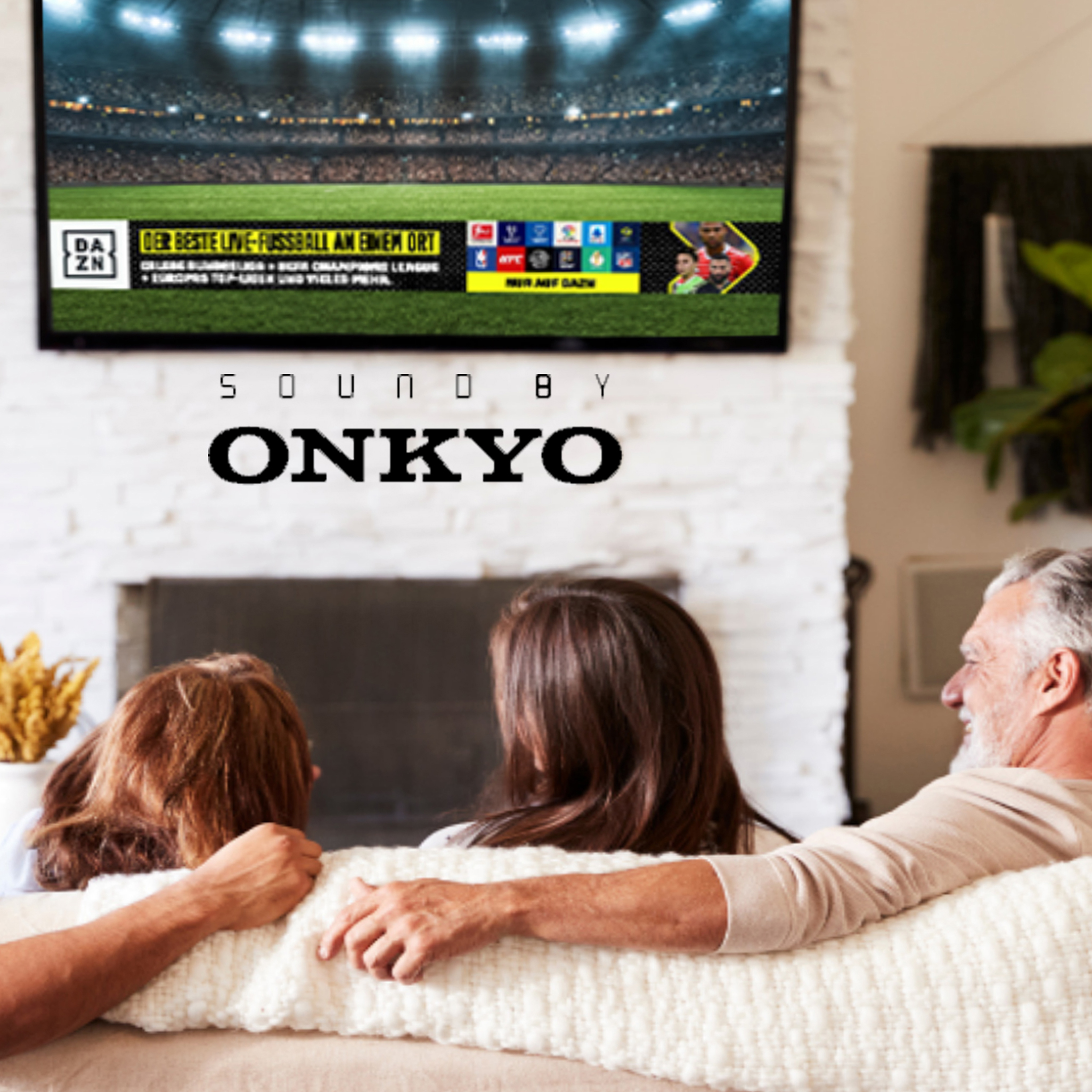 Familie schaut fern. DAZN-Logo auf dem Bildschirm mit Sport. Onkyo-Text darunter. Weißer Kamin und Deko.