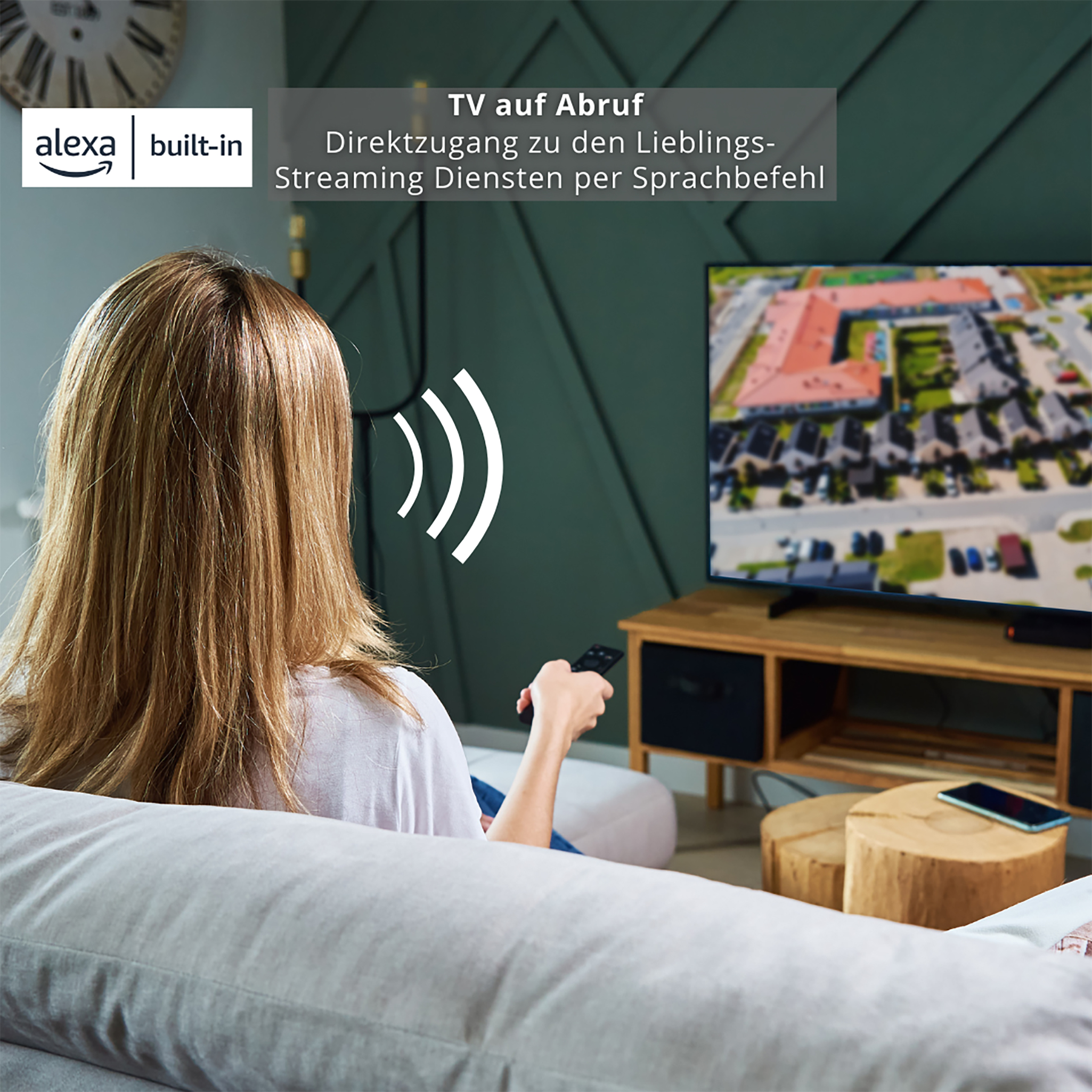 Frau mit Fernbedienung schaut fern. Alexa integriert. Fernseher auf Holzständer, Holzstühle.