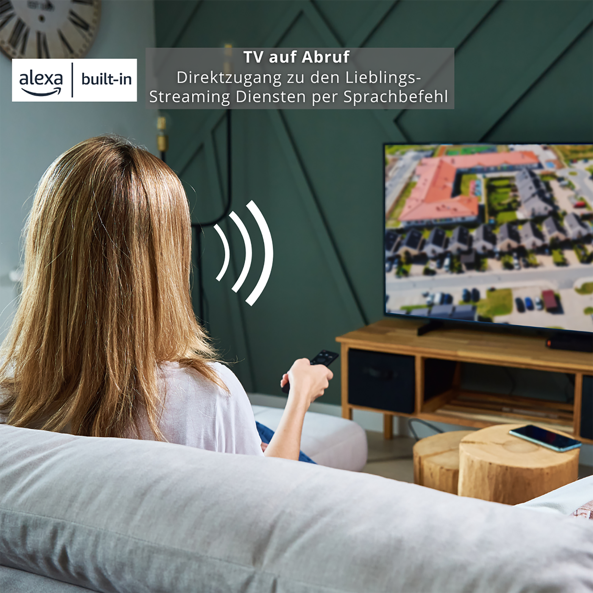 Frau mit Fernbedienung schaut fern. Alexa integriert. Fernseher auf Holzständer, Holzstühle.