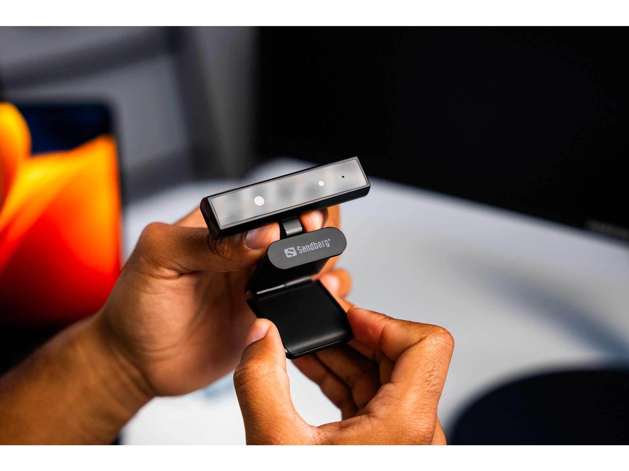 Ręce trzymają czarną kamerę internetową. Kamera ma klips do montażu na monitorze.