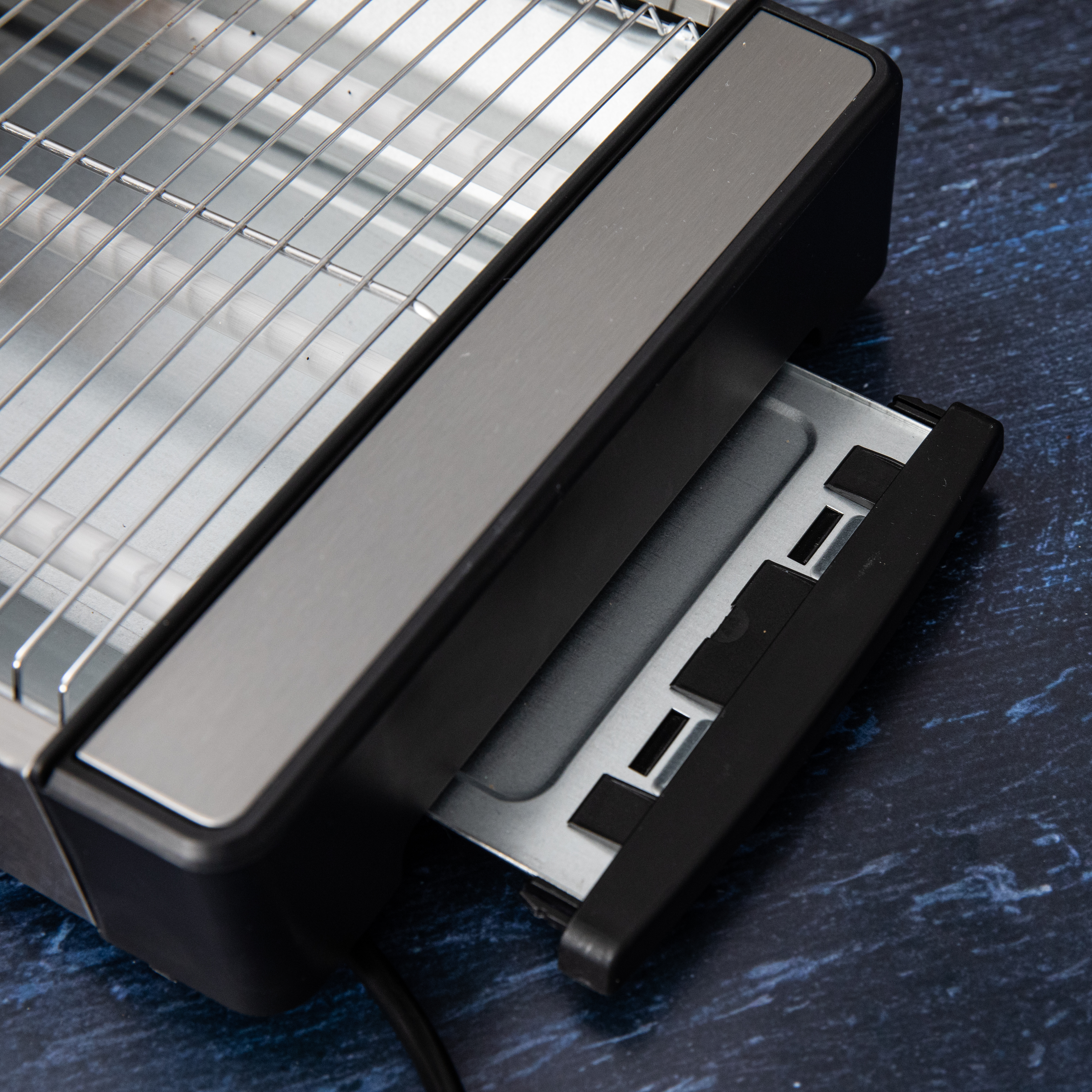 Zbliżenie tostera z otwartą tacką na okruchy. Taca jest czarna i błyszcząca.