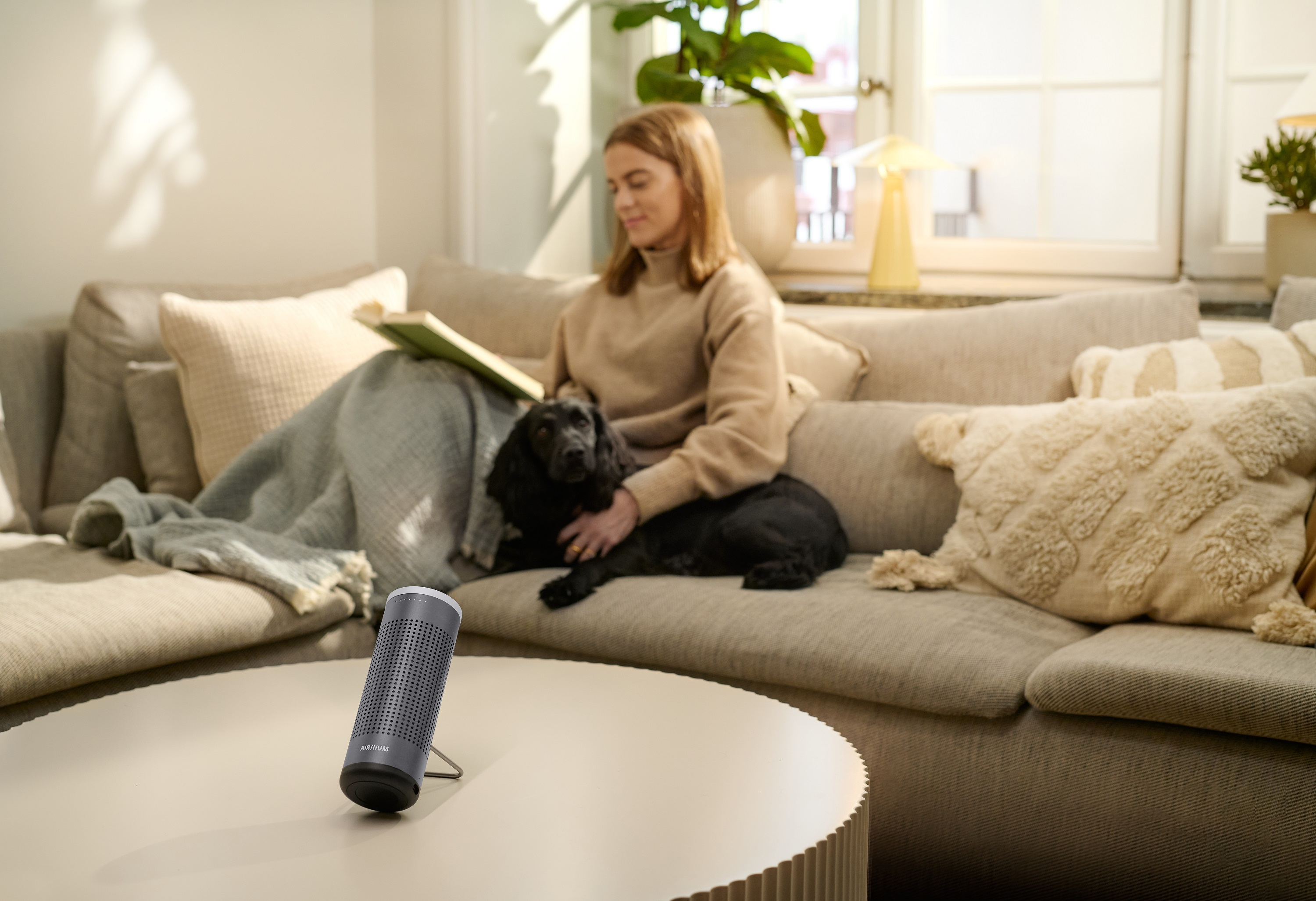 Frau mit Hund auf Sofa, Lautsprecher auf Tisch. Lesen eines Buches im Wohnzimmer.